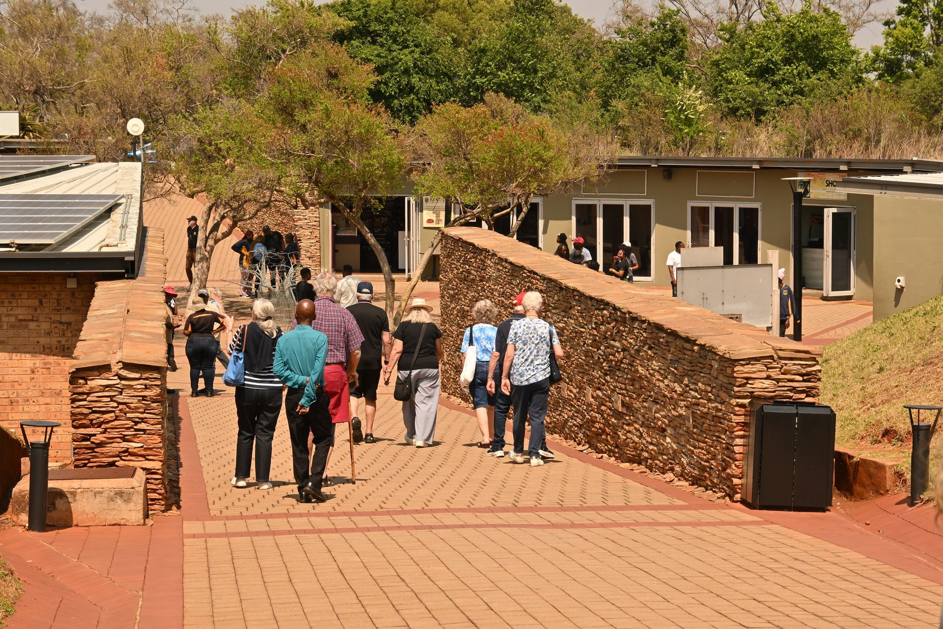 Group walking on a brick path between buildings in a sunny outdoor courtyard.