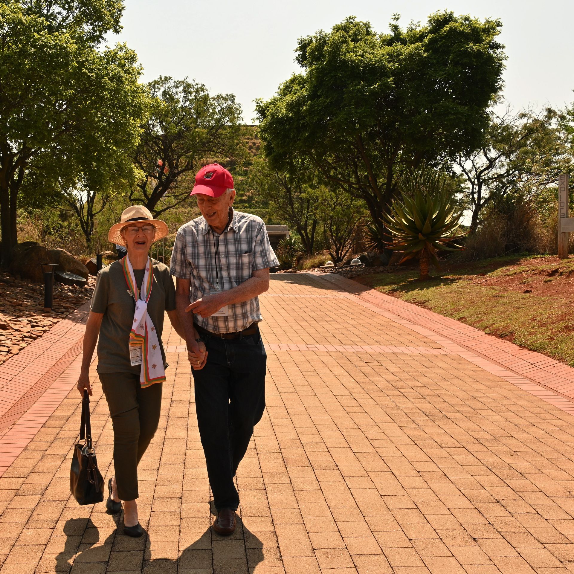 Two people walking on a sunlit brick path among trees, one in a red cap, one with a walking stick.