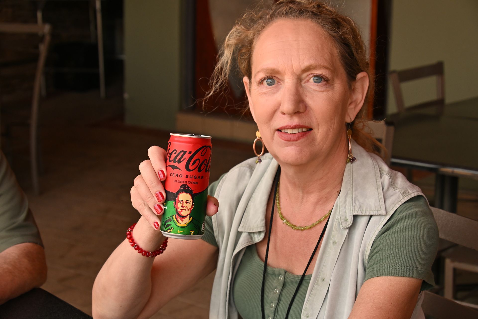 Woman holding up a Coca-Cola can, seated indoors at a table.