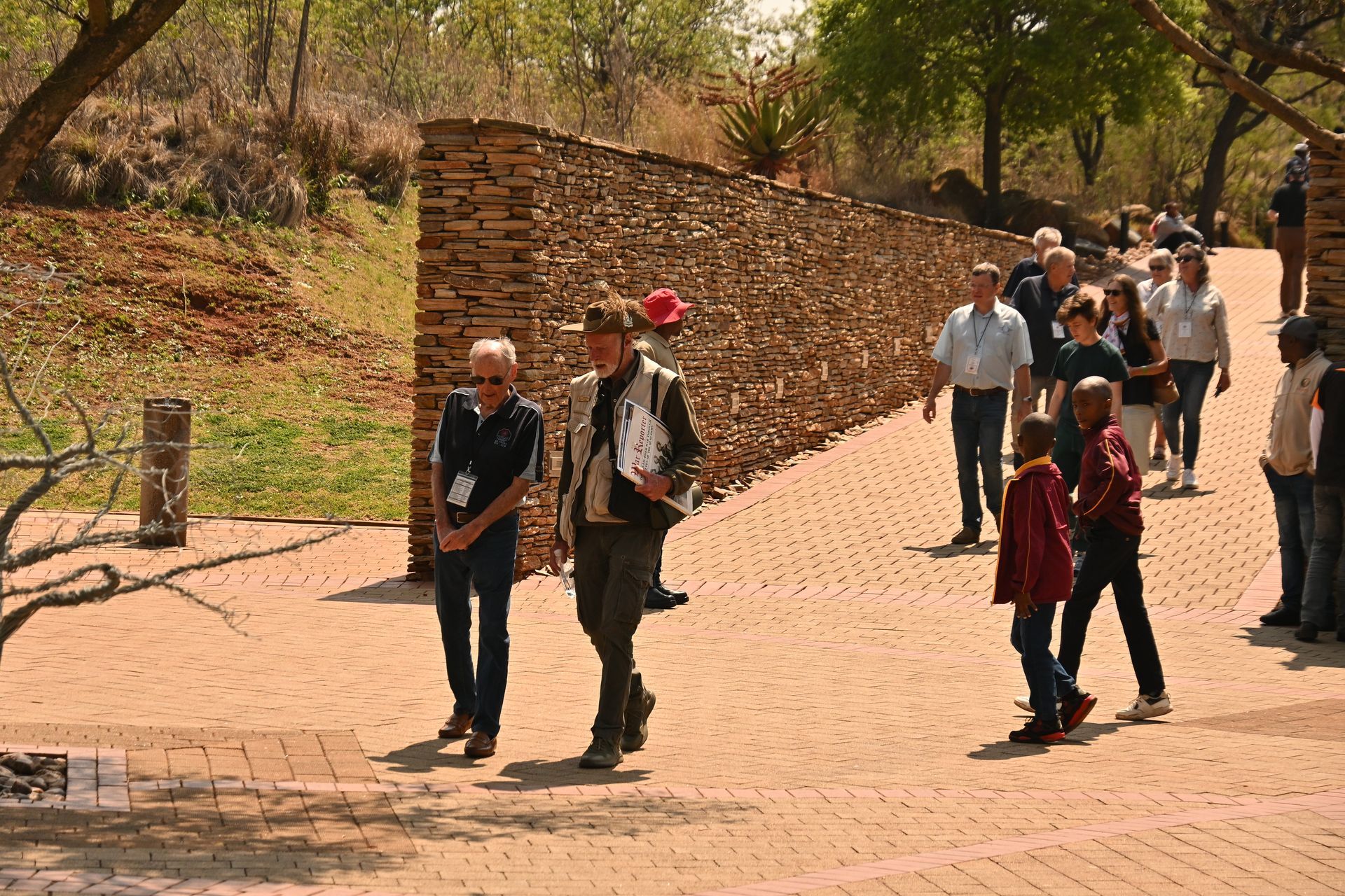 People walking along a sunny brick path beside a stone wall and trees