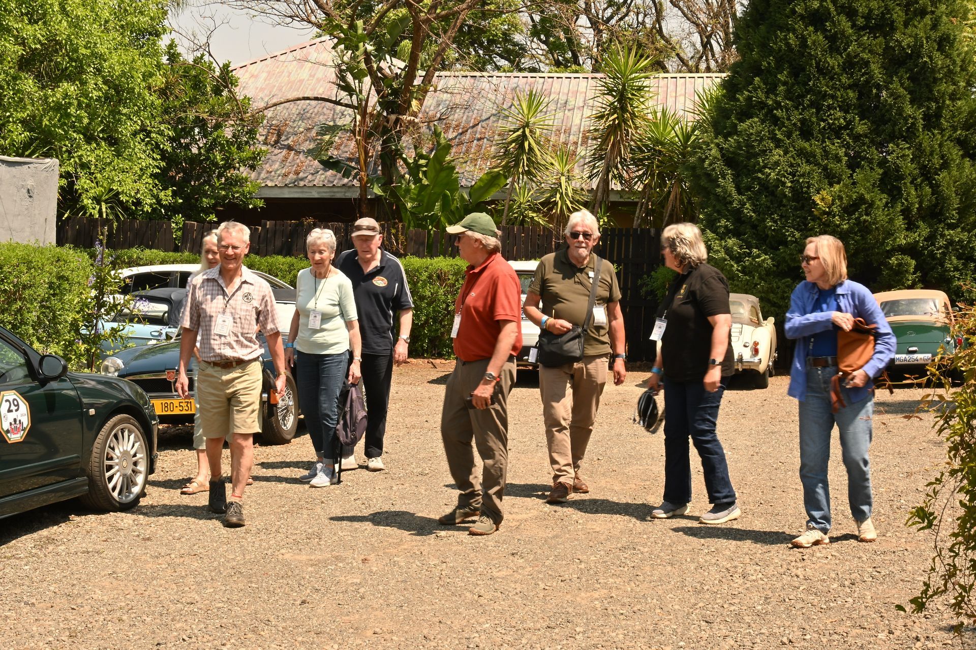 Group of people standing and talking beside parked cars on a gravel driveway in bright sunlight
