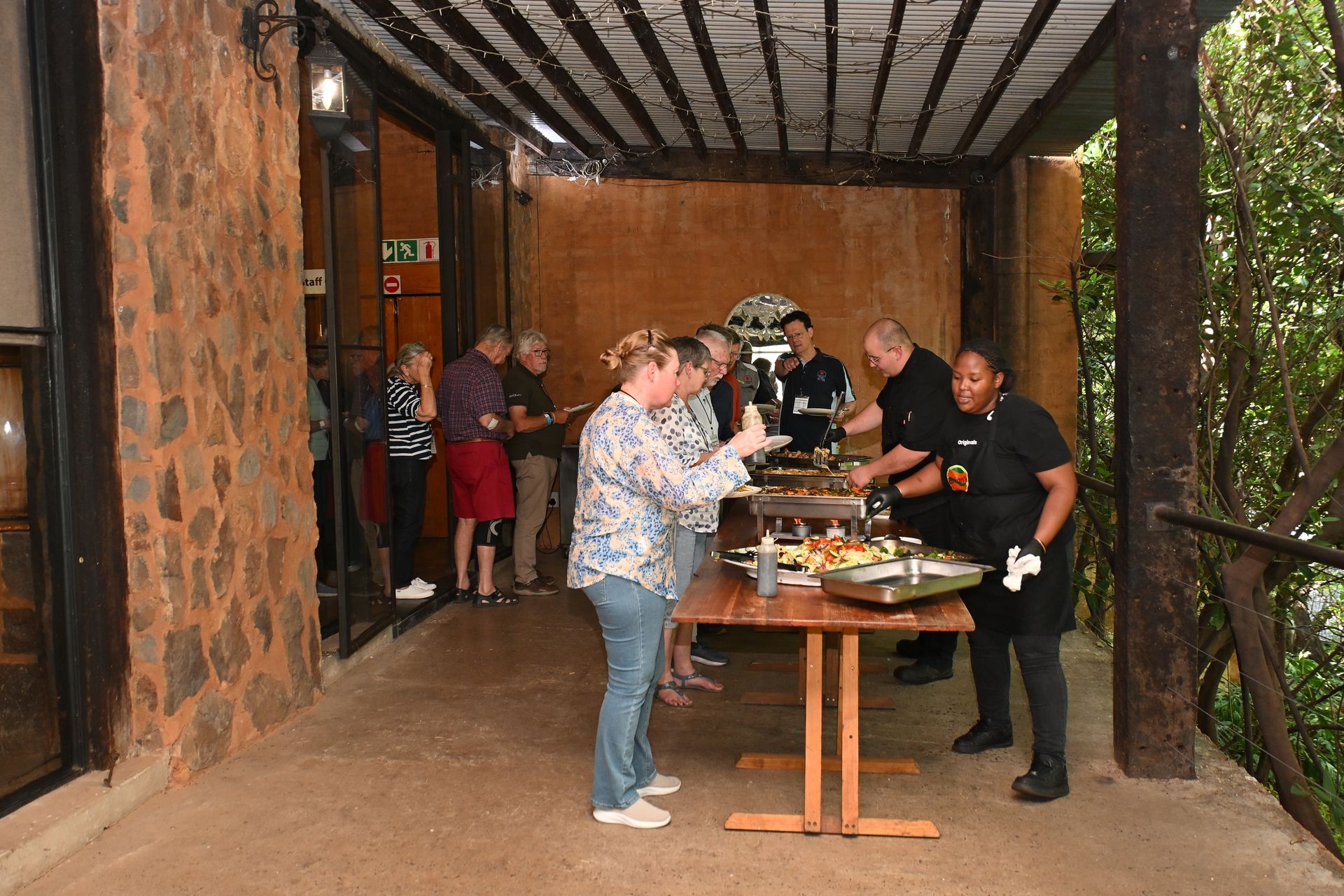 People serving food at a long table in a covered outdoor walkway with brick walls and greenery nearby