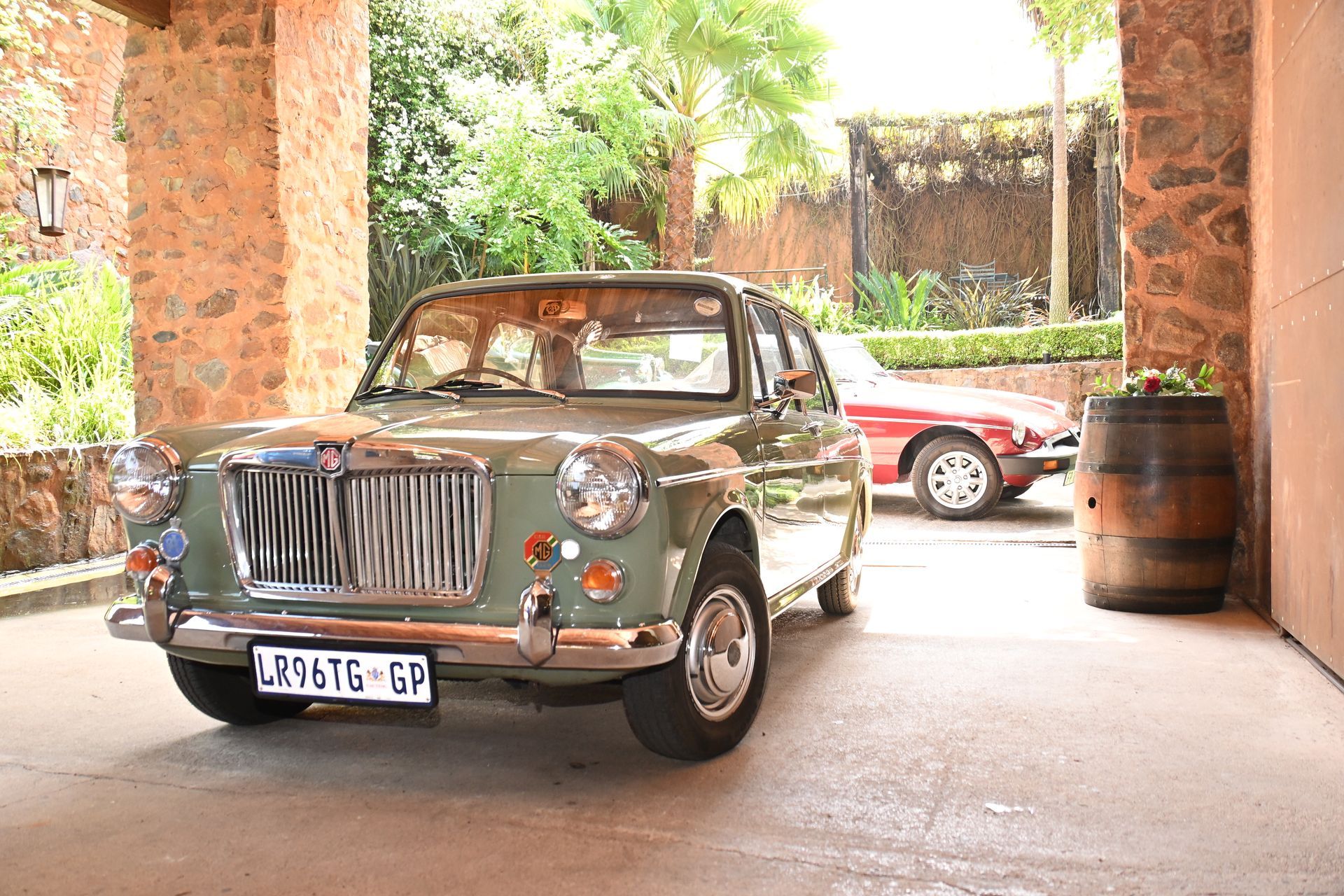 Vintage green car parked under a covered driveway with a red car and wooden barrel nearby