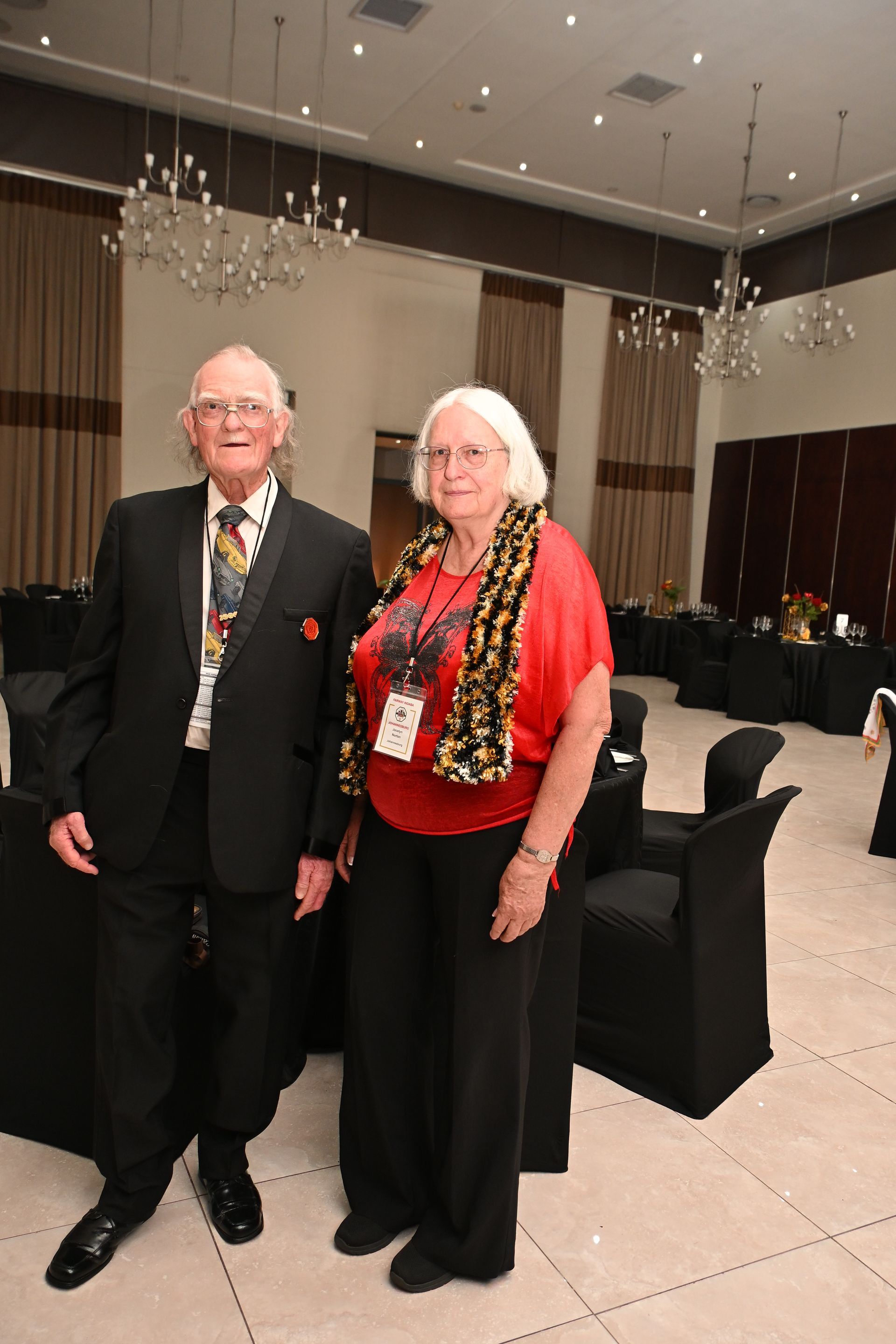 Two people in formal wear standing in a banquet hall with black table settings and chandeliers.