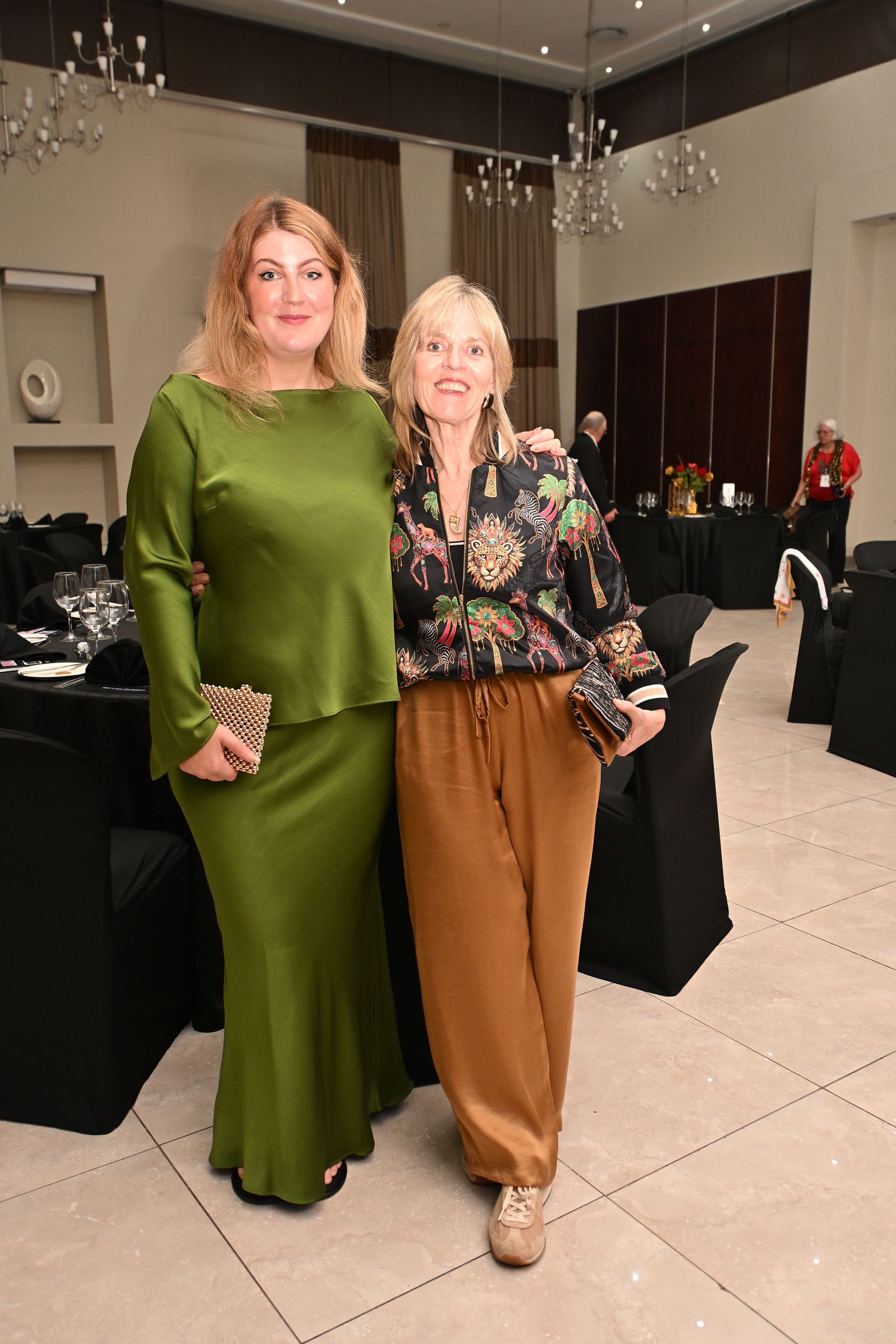 Two women posing at a formal event in a banquet hall, one in green and one in floral blouse with brown pants.