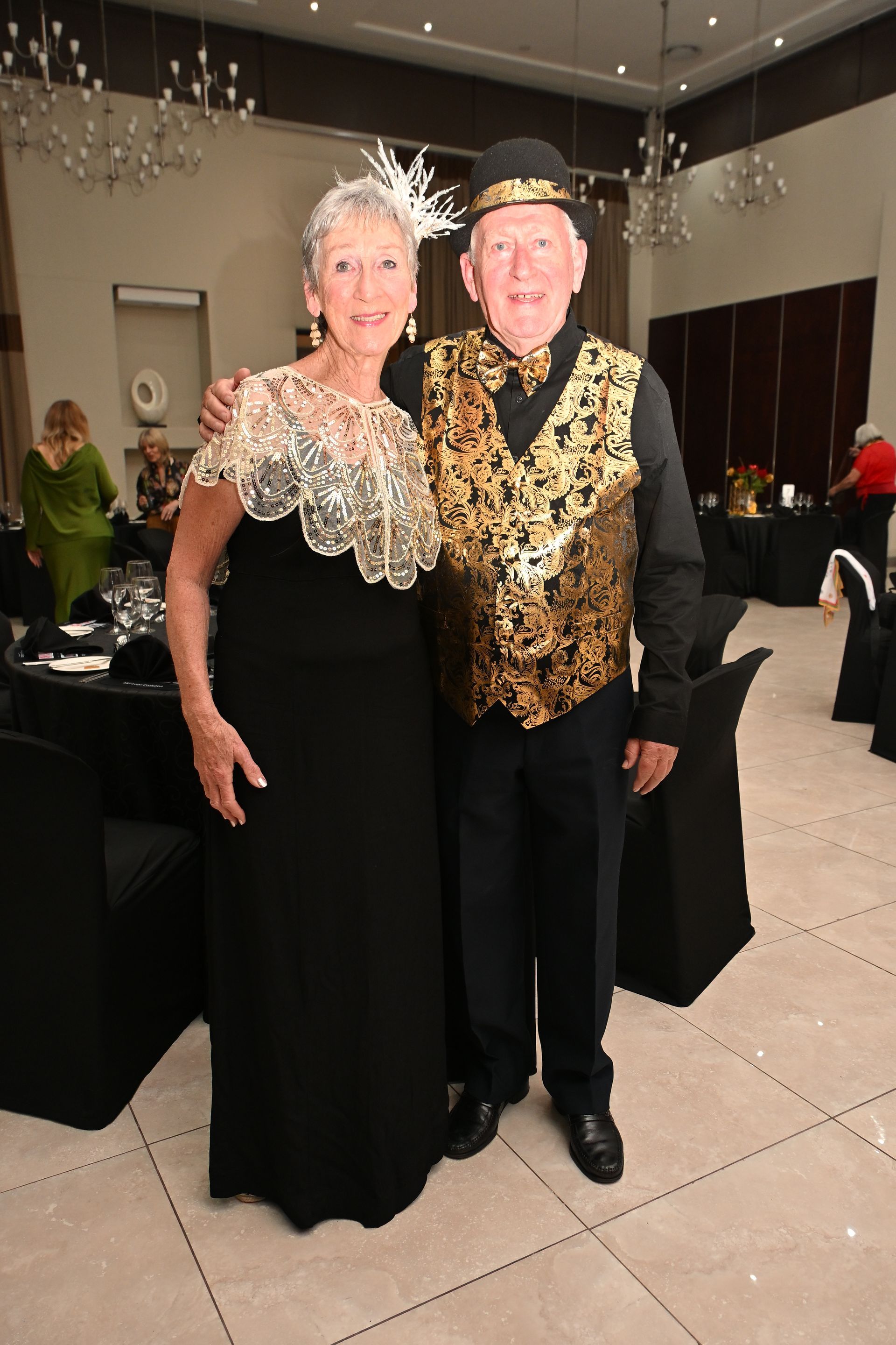 Two people posing in formal black-and-gold outfits at an indoor event.