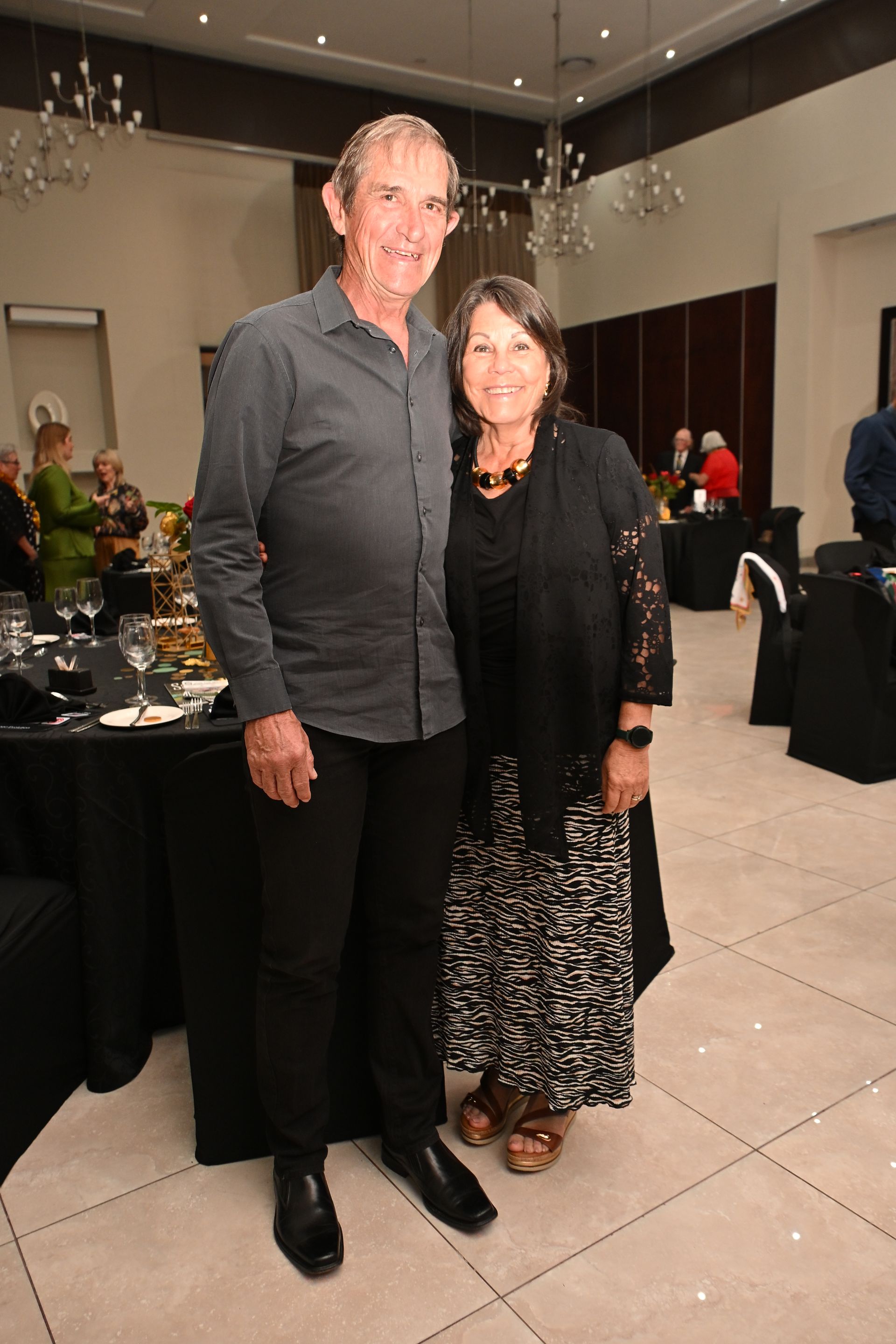 Two people posing together at a formal indoor event, with tables and guests in the background.
