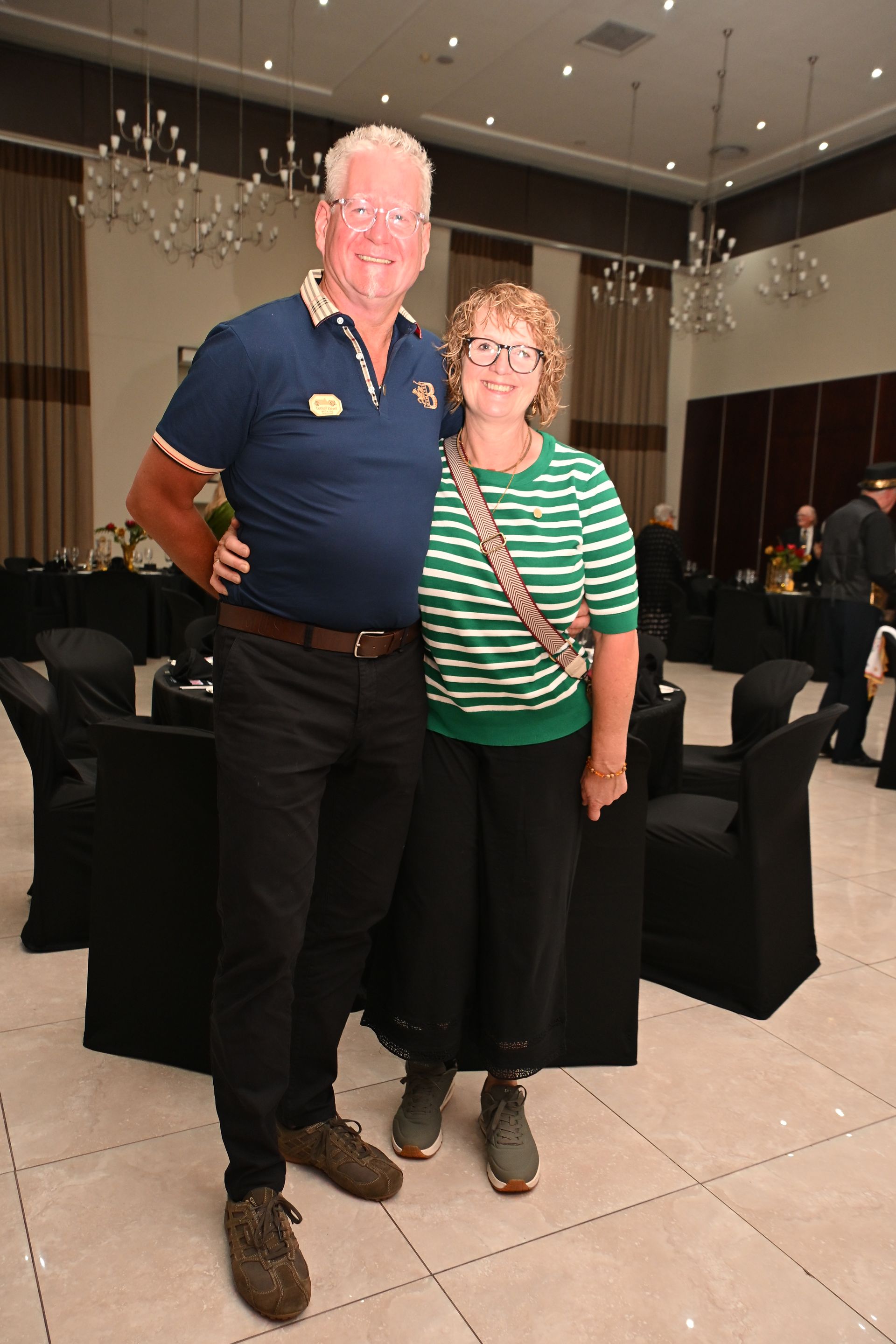 Two people posing together in a banquet hall with black chairs and hanging lights