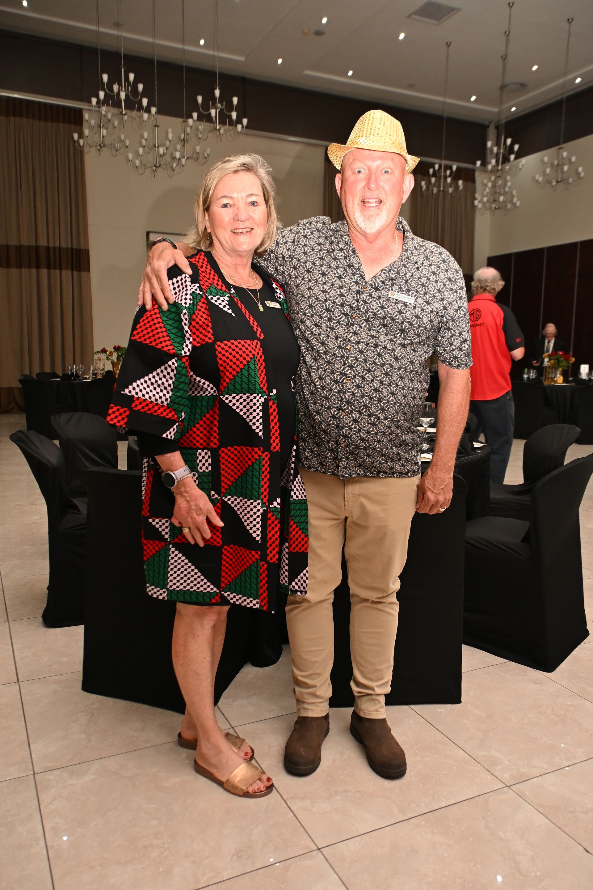 Two people smiling at an indoor event, standing together in front of black-covered tables.