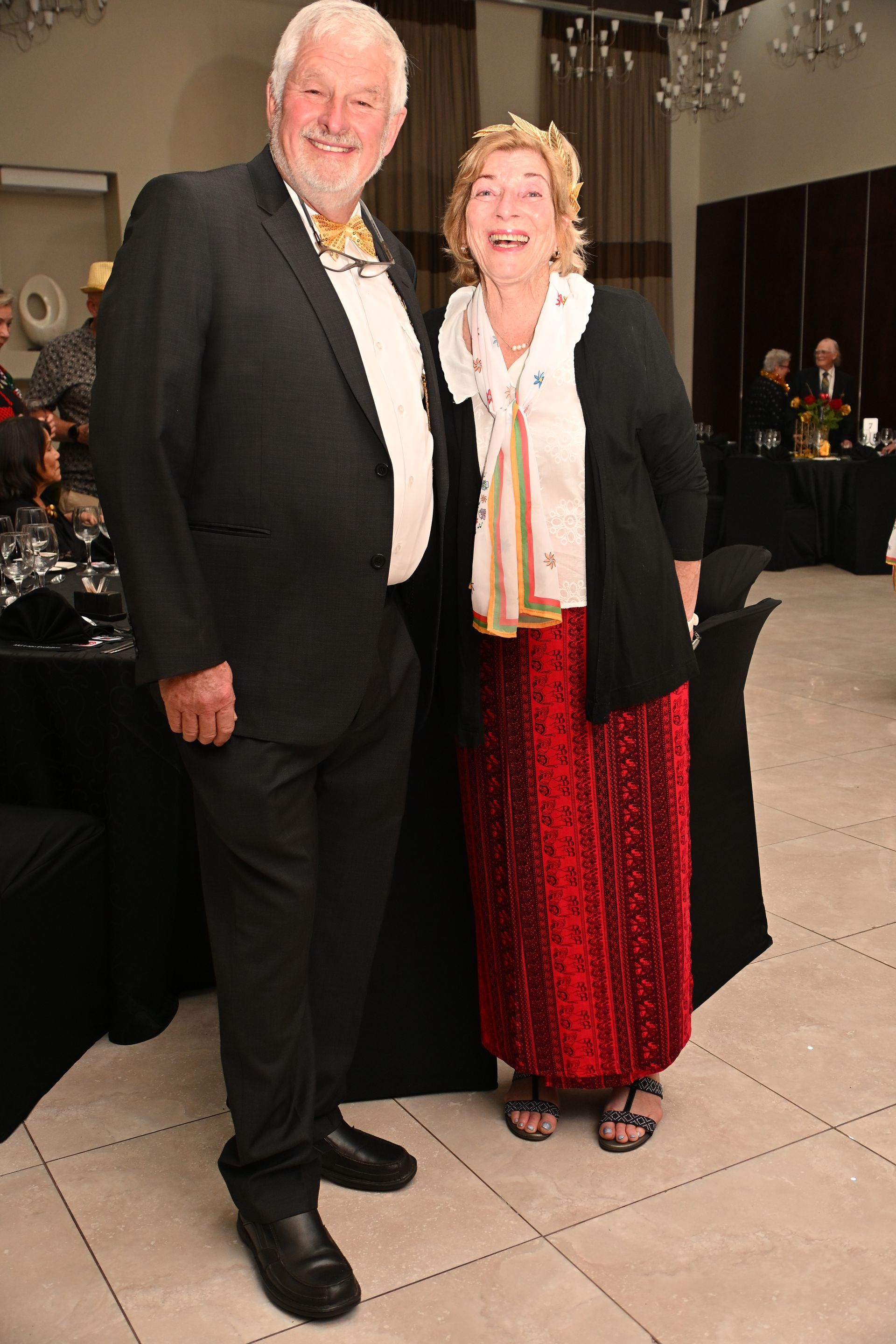 Two people in formal attire posing together at an indoor event, smiling at the camera.