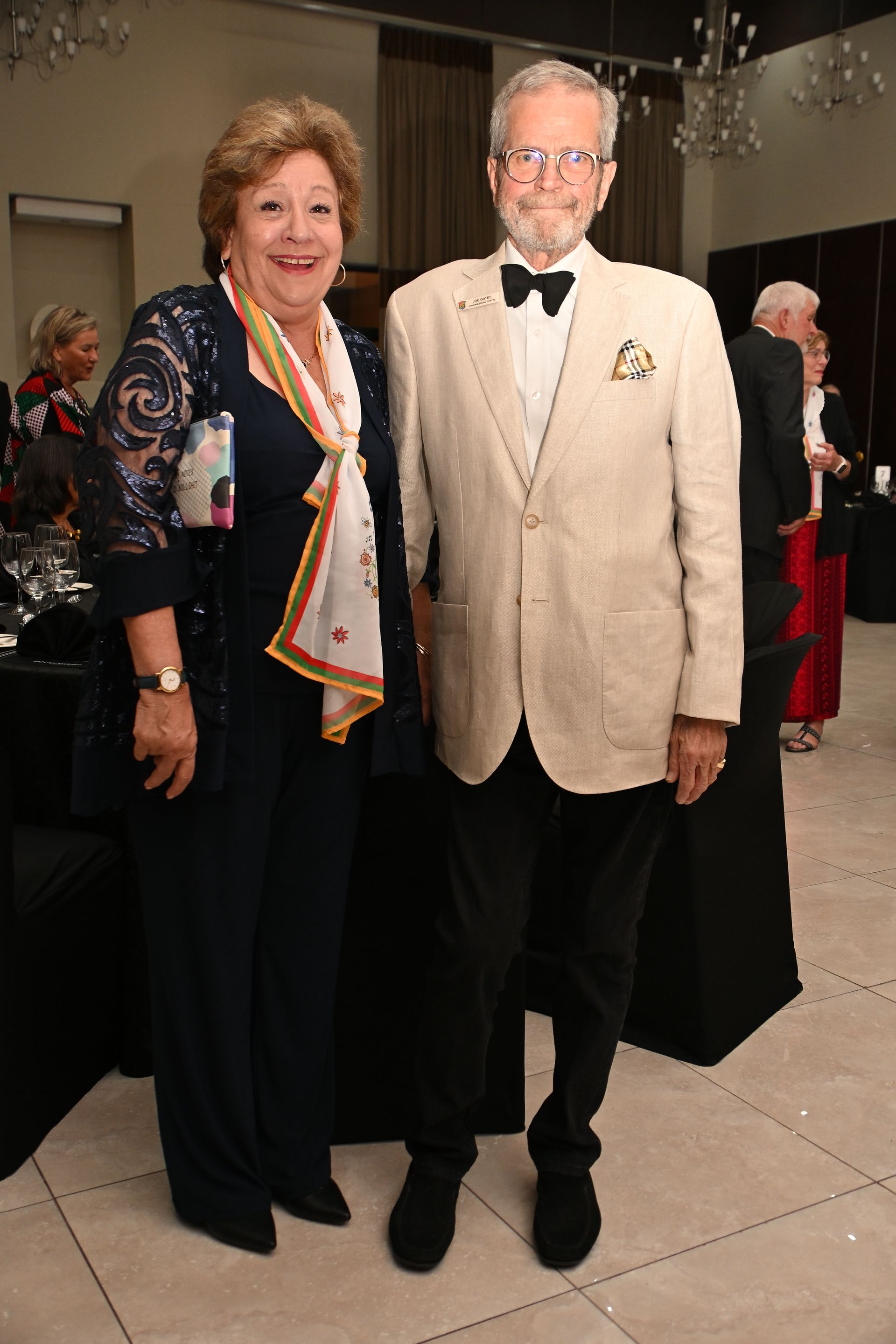 Two people posing at a formal event, wearing evening attire, with curtains and other guests in the background