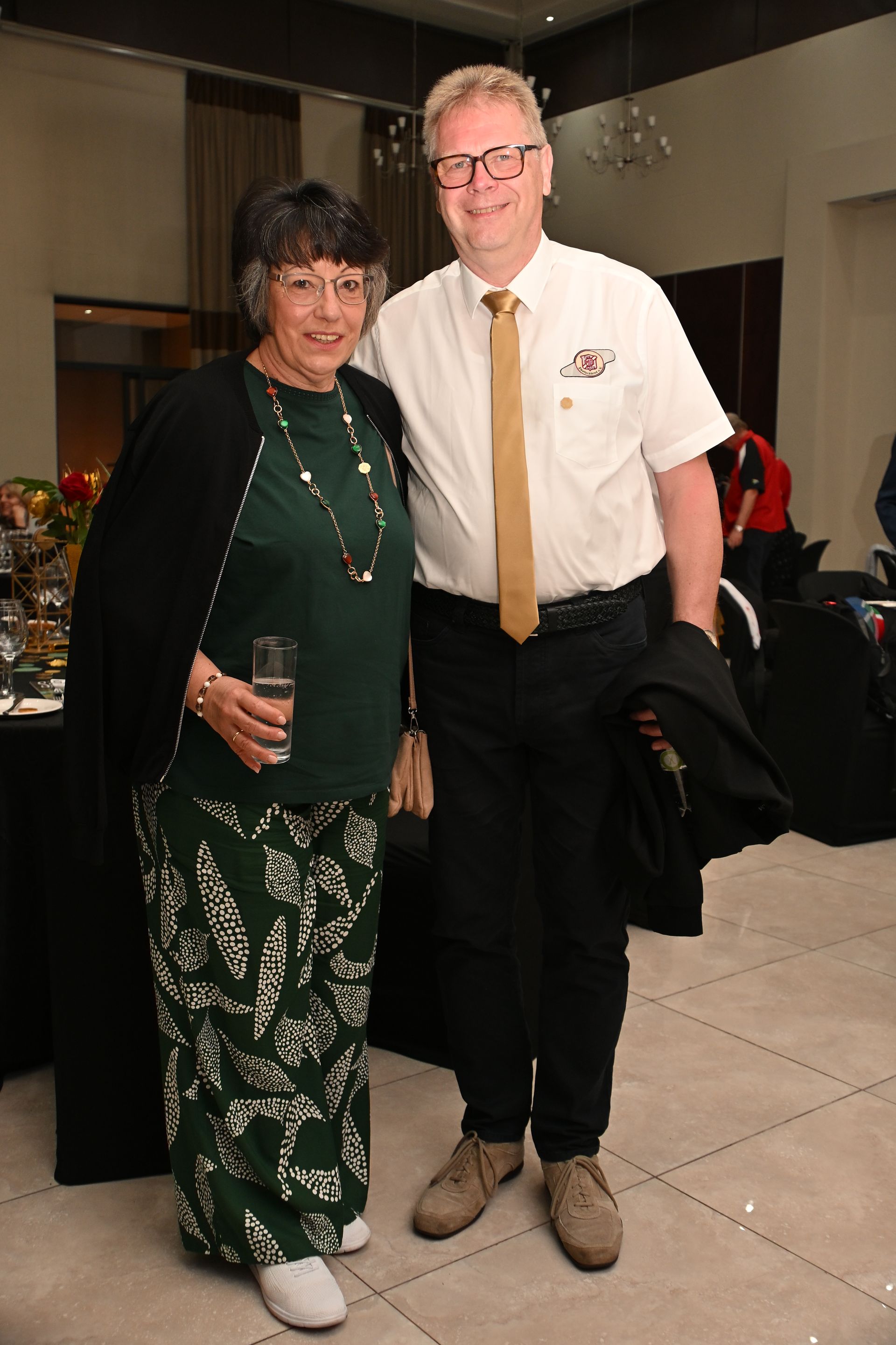 Two people posing indoors, one in a green dress holding a drink, the other in a white shirt and tie.