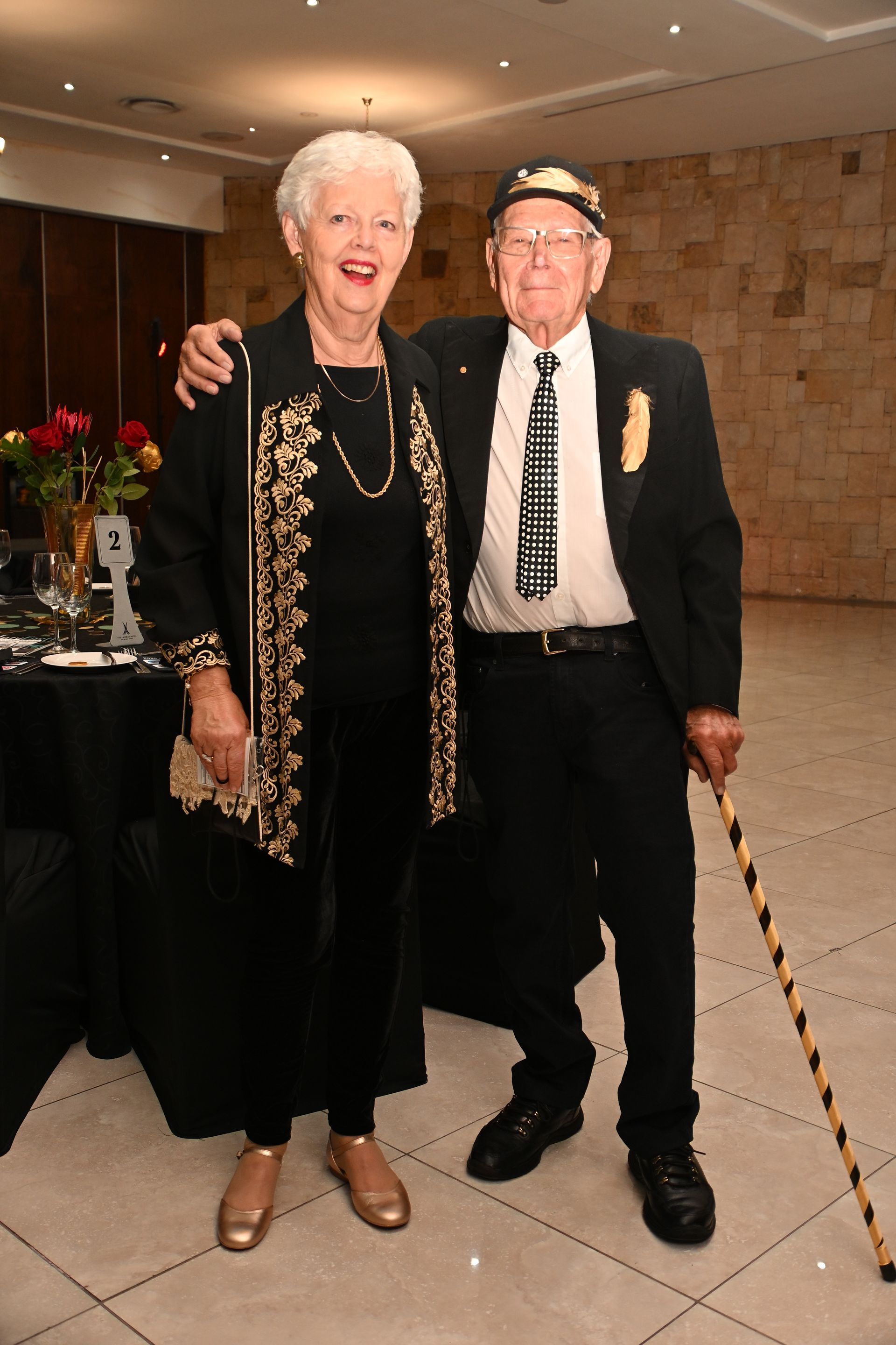 Two people posing together at an indoor event, one holding a cane and both dressed formally.