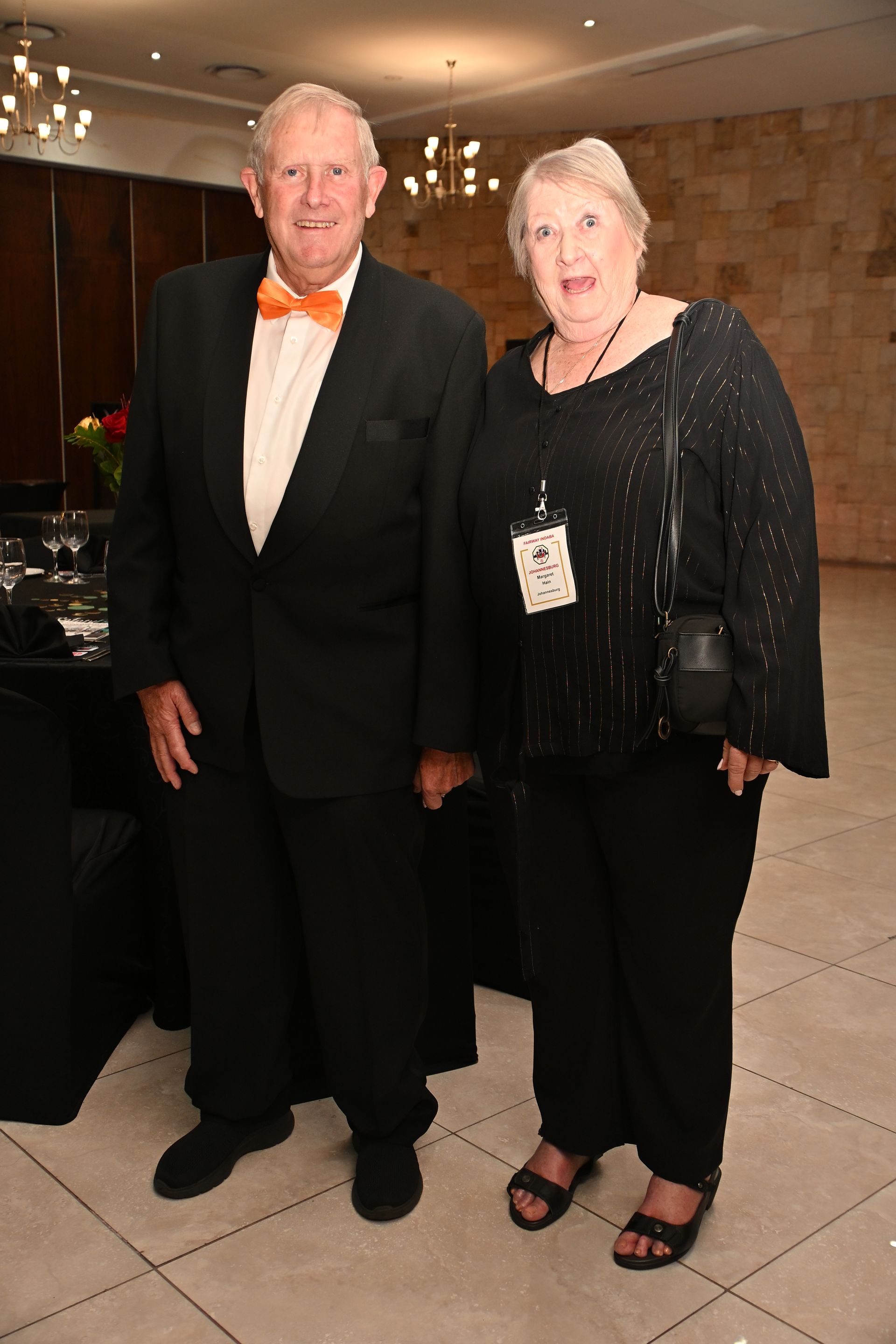 Two people posing indoors at a formal event, dressed in black with an orange bow tie and red shoes.