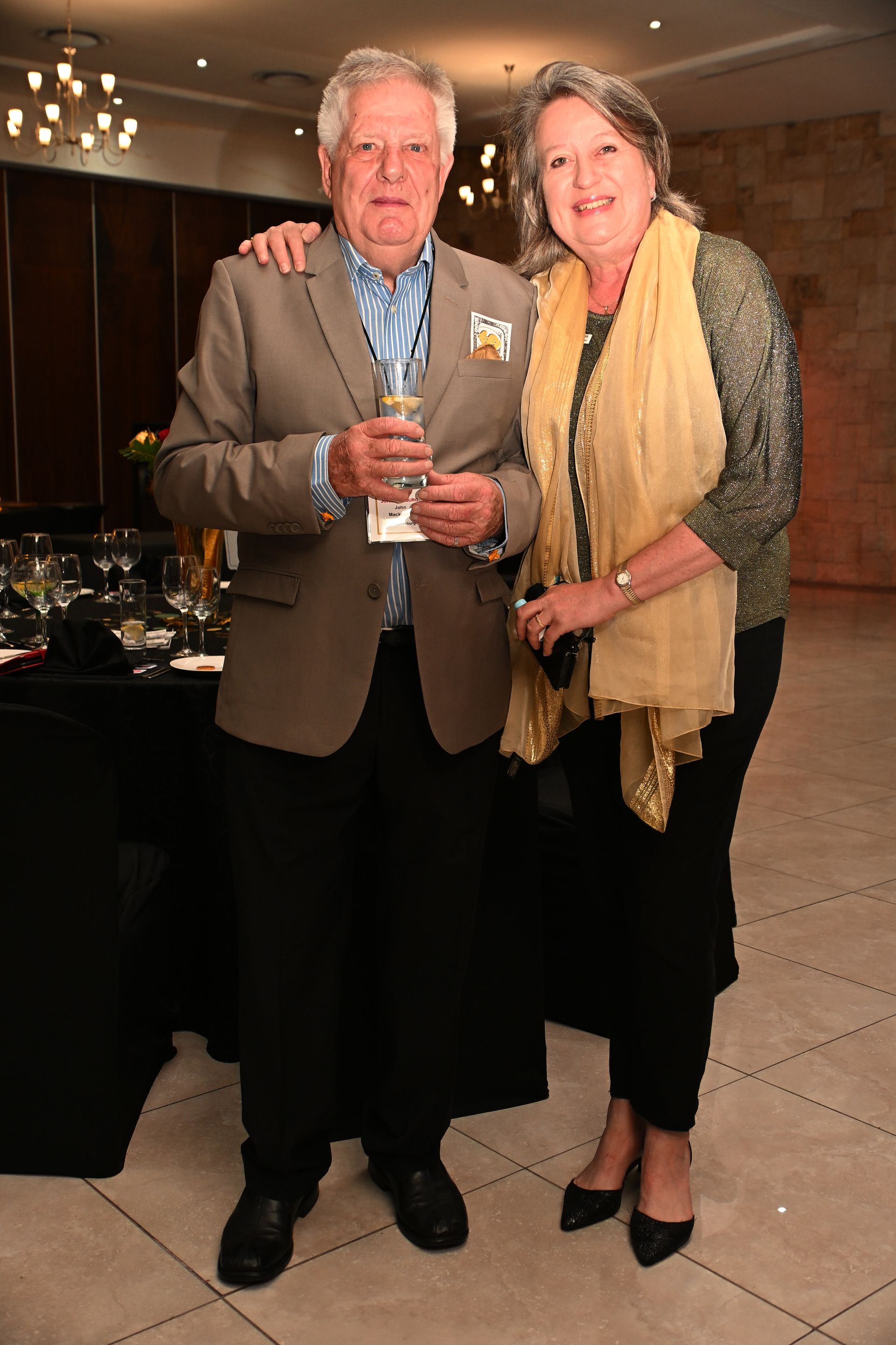 Two people standing indoors at a party, holding drinks and smiling in formal attire.