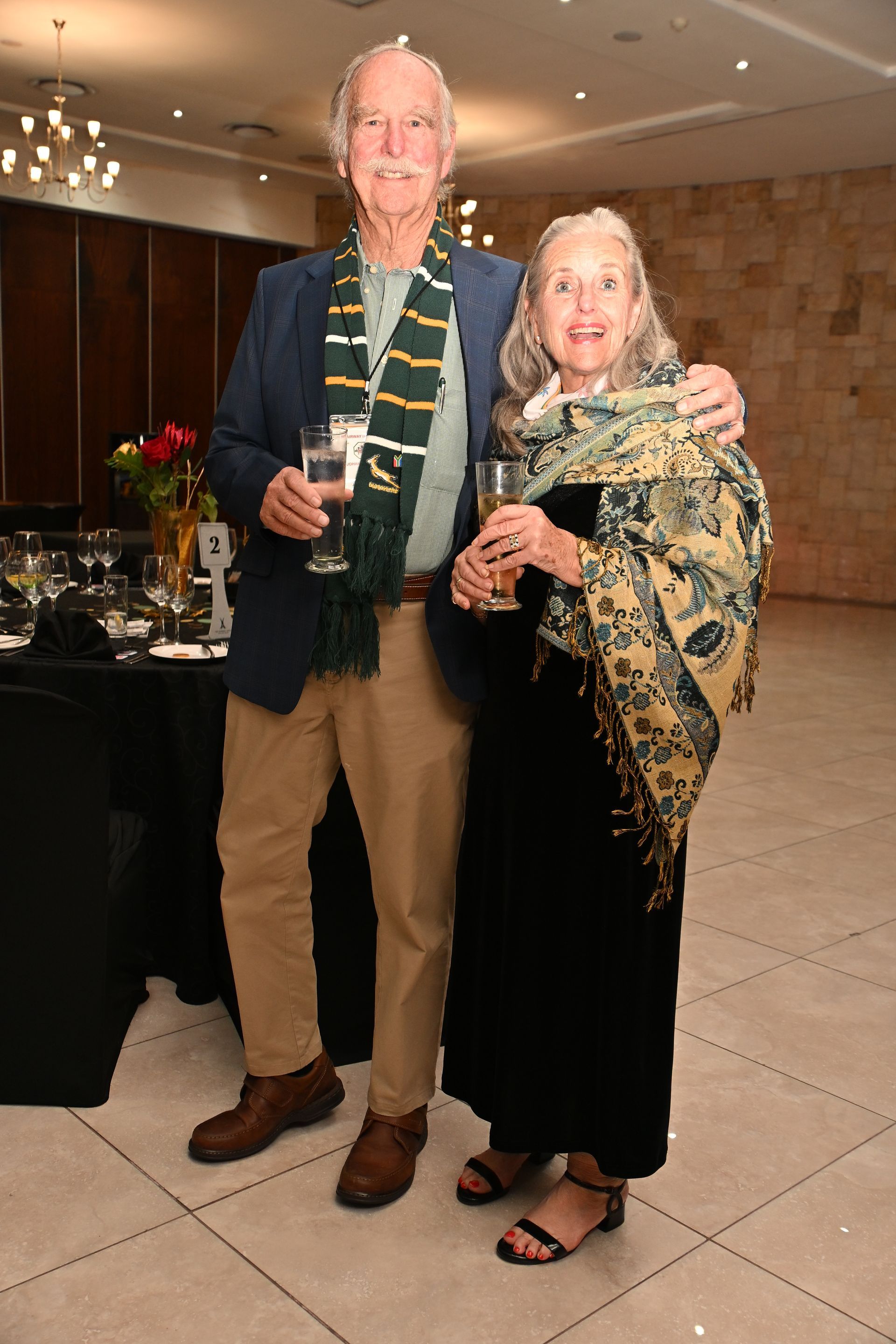 Two people posing with drinks in a warmly lit indoor event hall, dressed in formal attire.