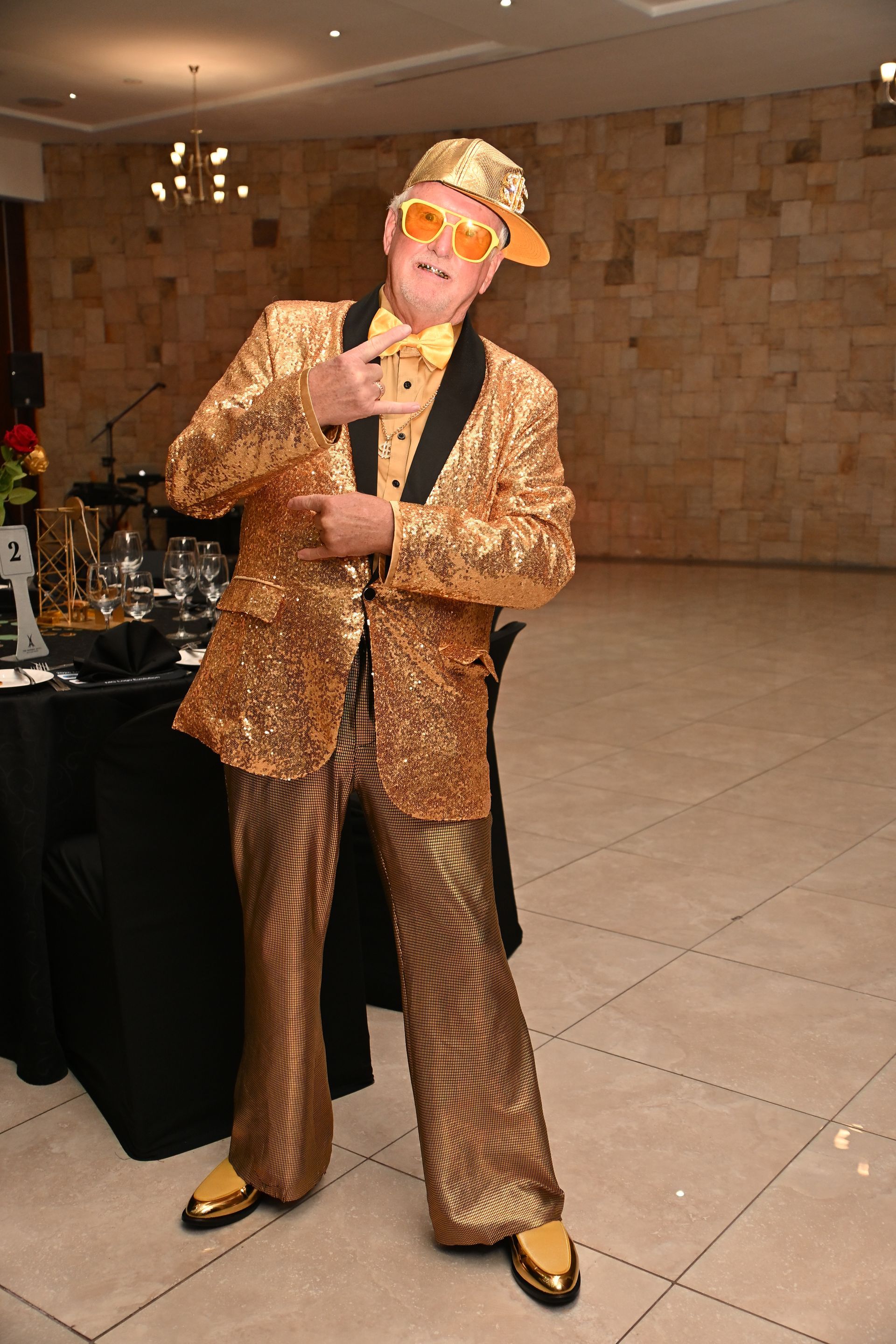 Person in a gold sequin suit and hat posing indoors at an event hall.