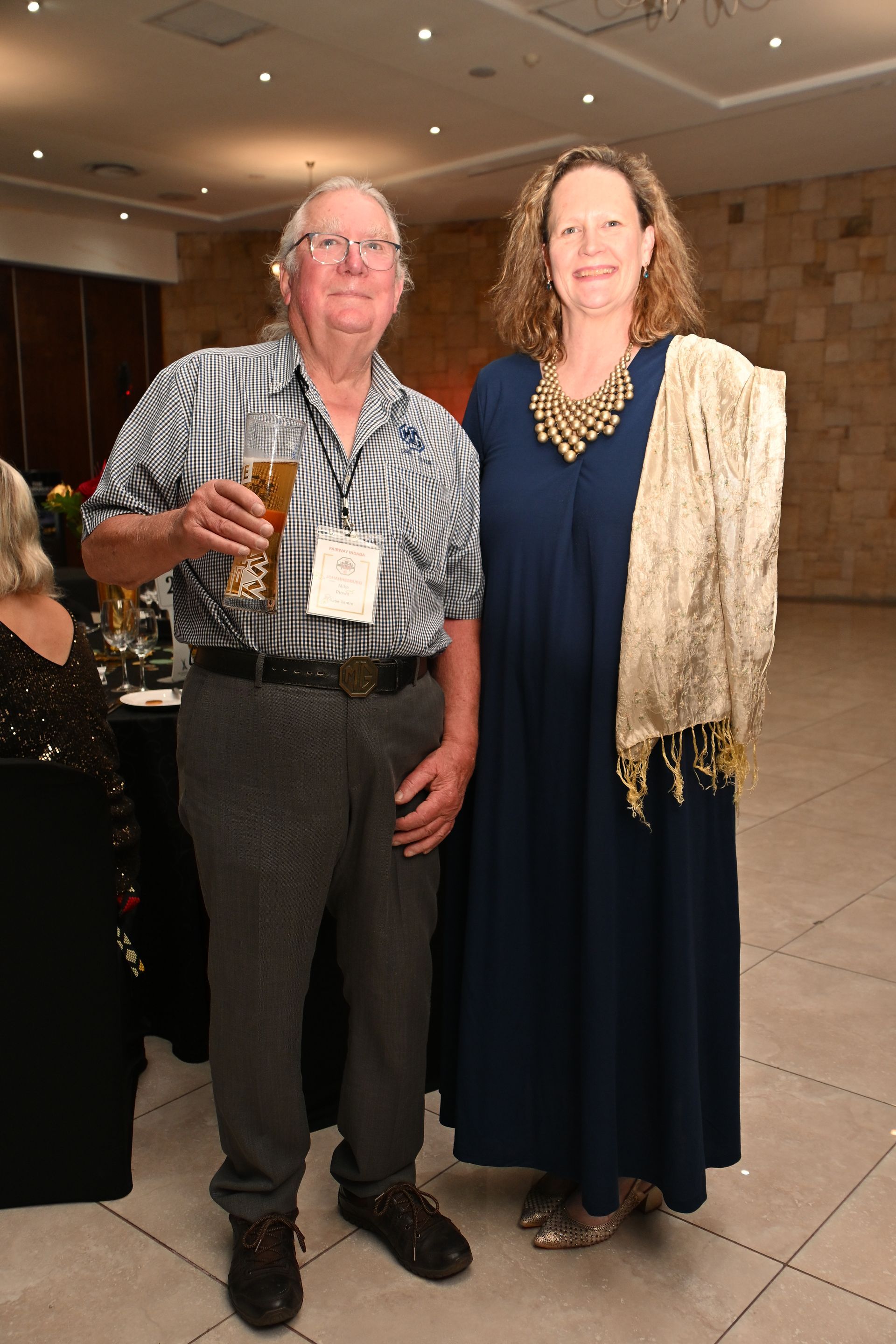 Two people posing indoors at a formal event, one holding a drink and the other wearing a navy dress and gold shawl