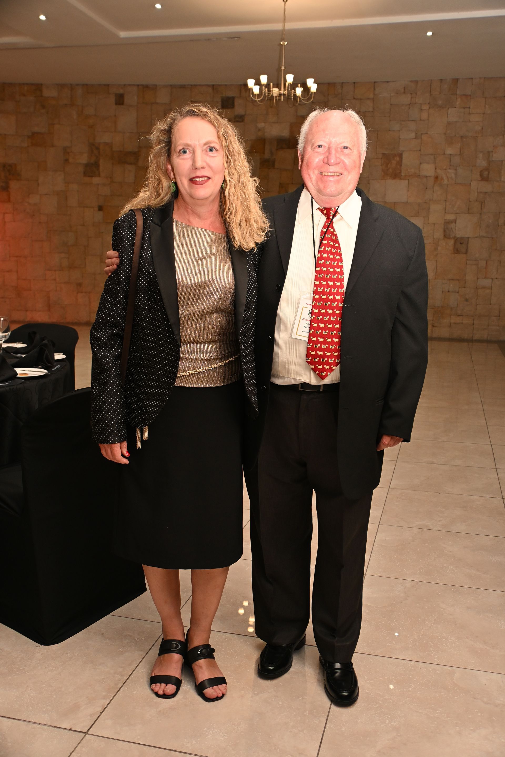 Two people posing indoors, wearing formal attire, in a warmly lit room.