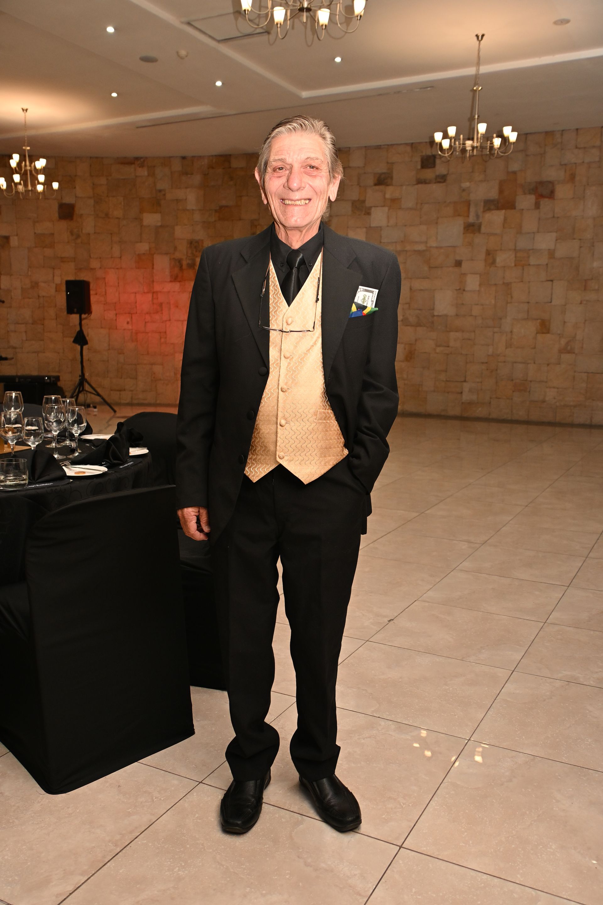 Man in black suit and gold vest standing in a banquet hall with chandeliers and tiled floor