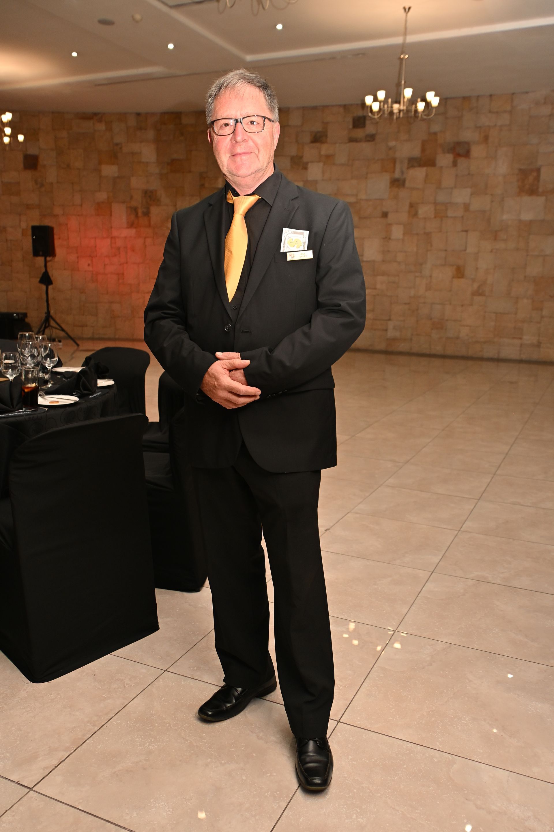 Man in a black suit with a gold tie standing at an event venue beside a table