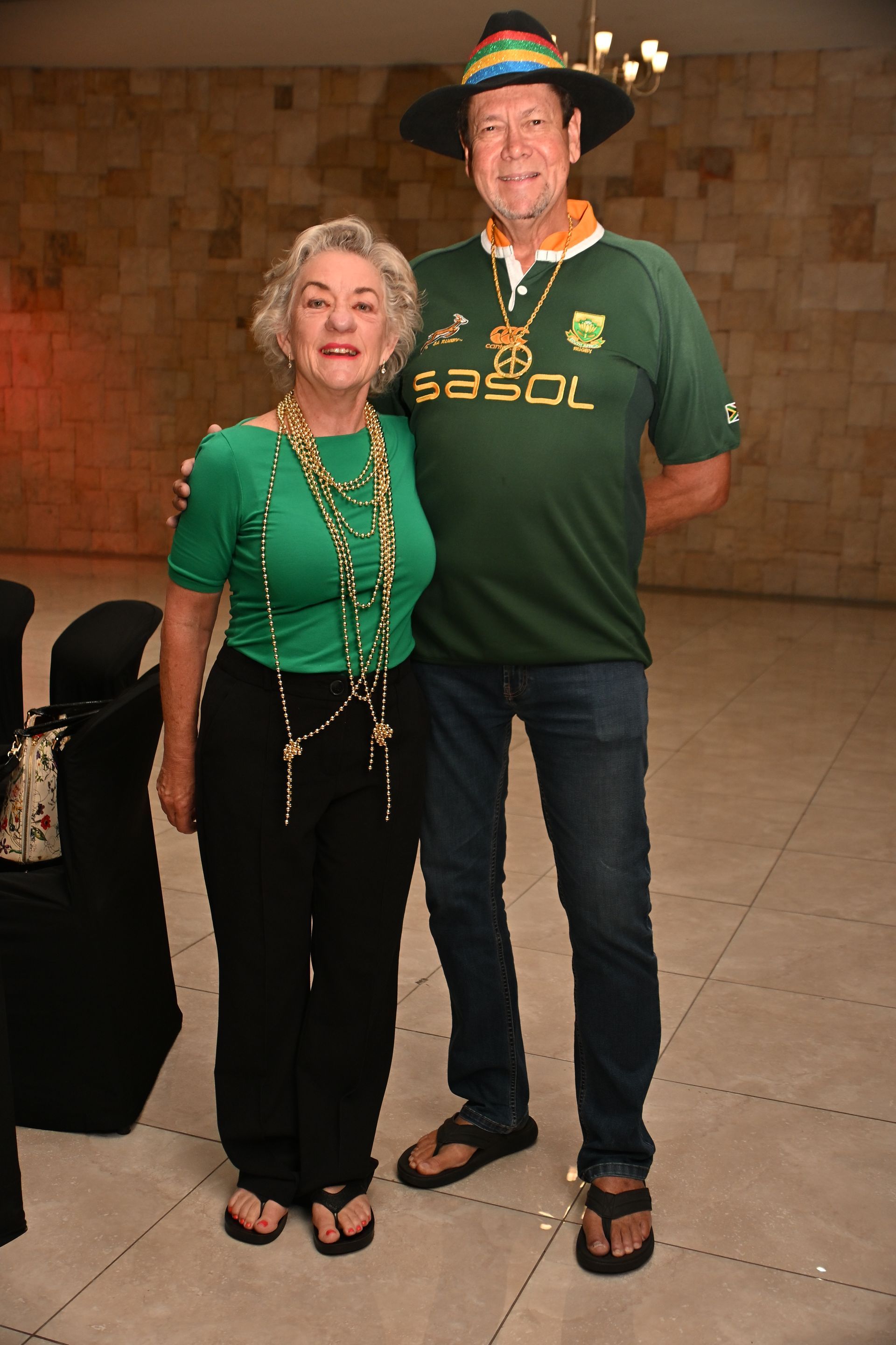 Two people in green shirts stand together indoors, smiling beside a suitcase.