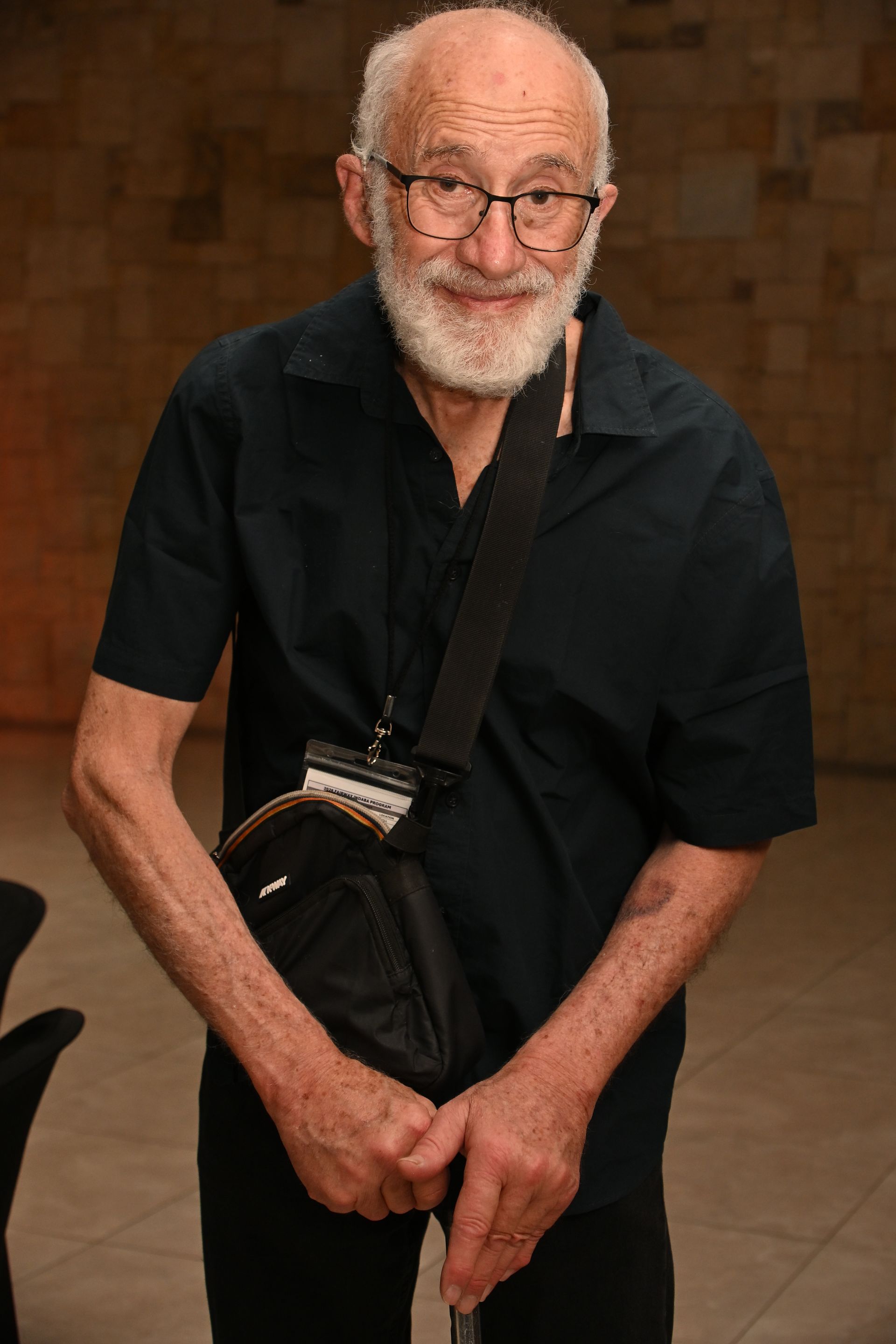 Smiling man in black shirt and glasses stands indoors with a shoulder bag and lanyard