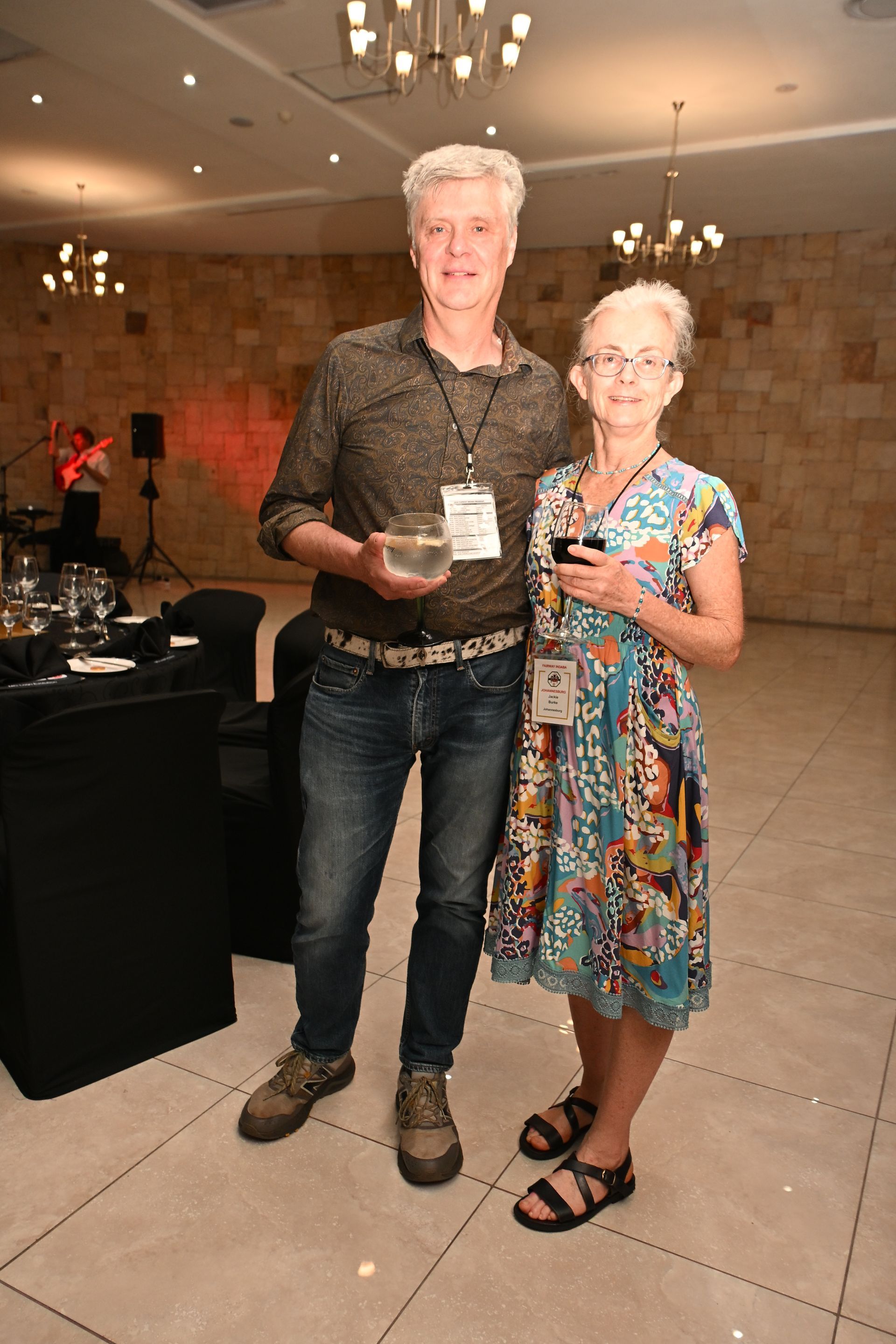 Two people standing together at an indoor event, one holding a drink and the other in a floral dress.