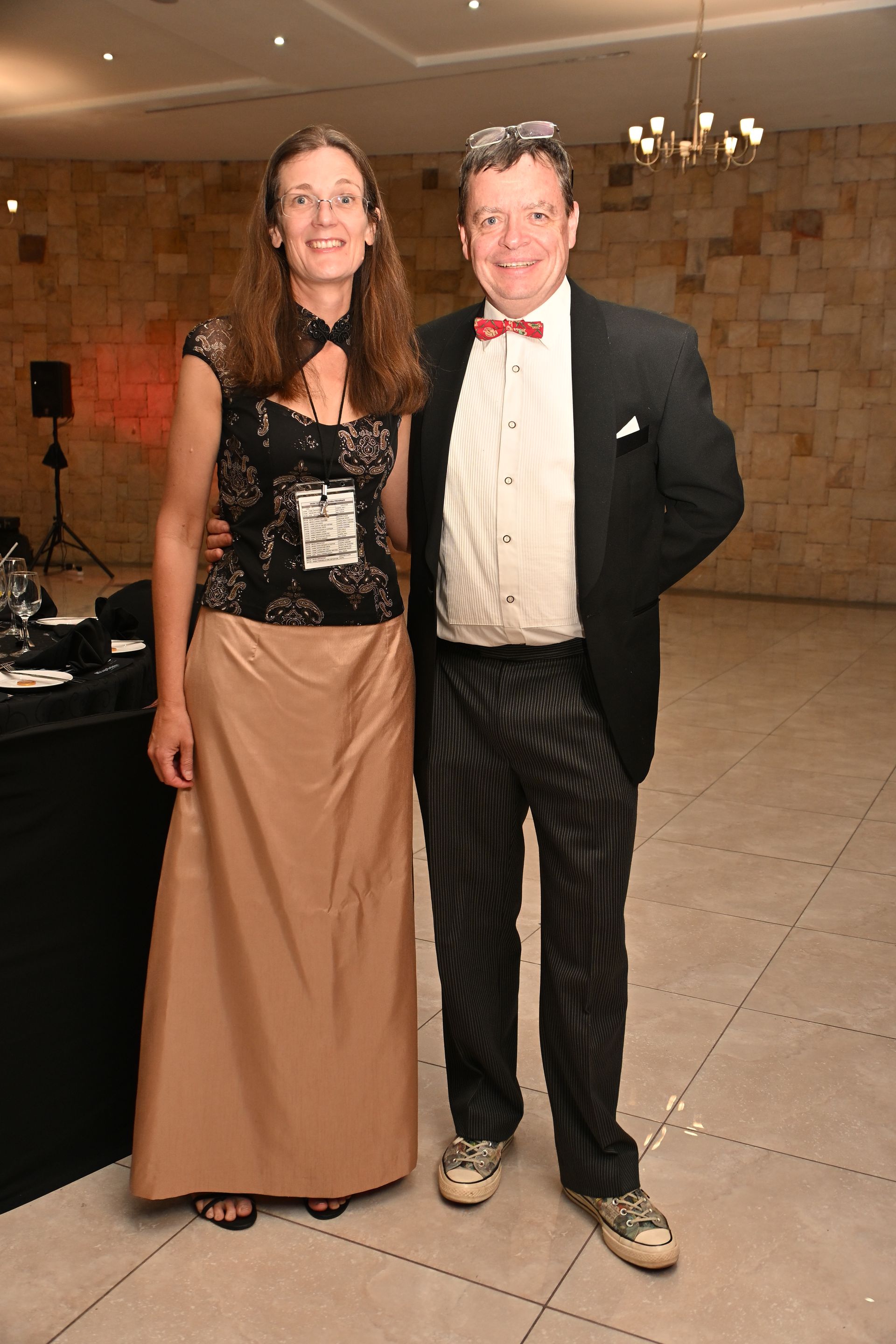 Two people posing together at a formal indoor event, wearing dressy black and tan outfits.