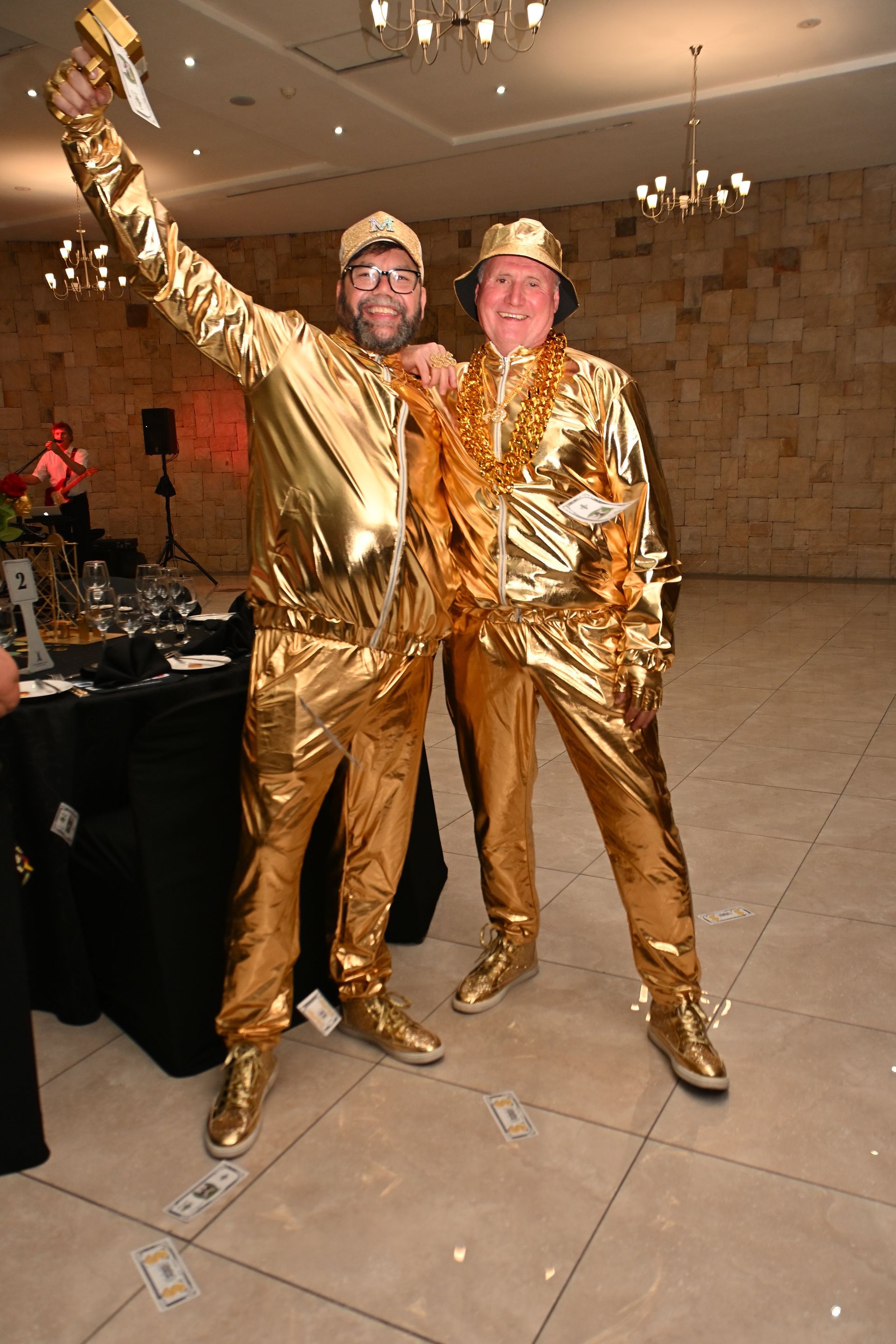 Two people in shiny gold outfits posing and smiling in a banquet hall, one raising an arm.