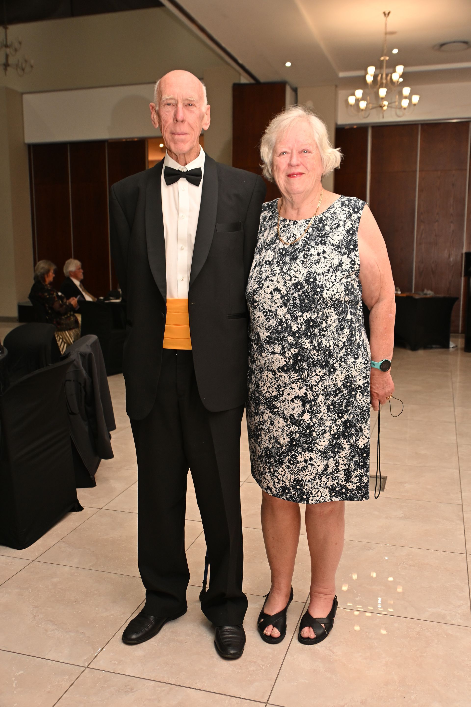 Two people in formalwear standing indoors at a formal event, with tables and chandeliers in the background.