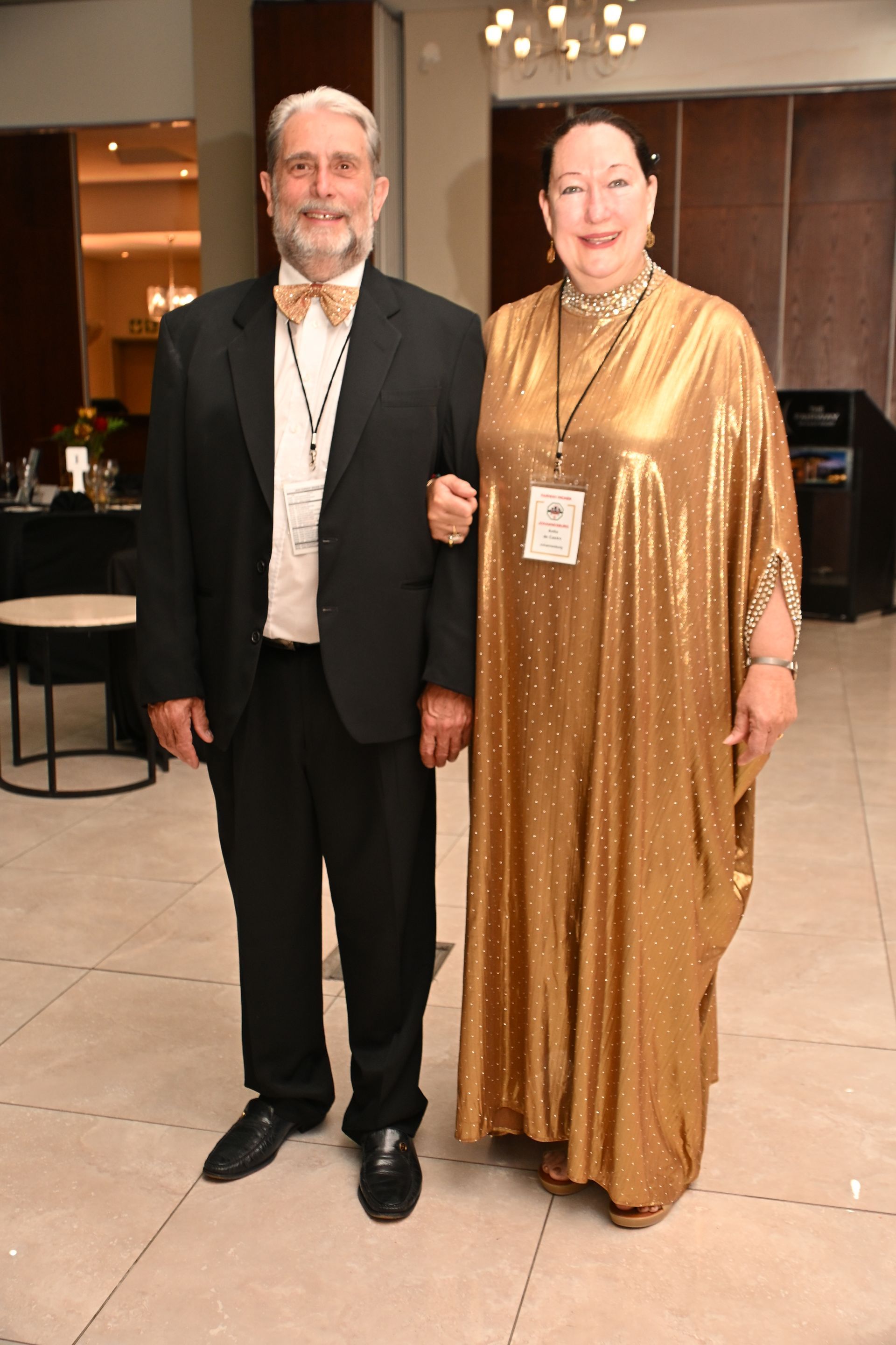 Two people posing indoors at a formal event, one in a black suit and one in a gold gown.