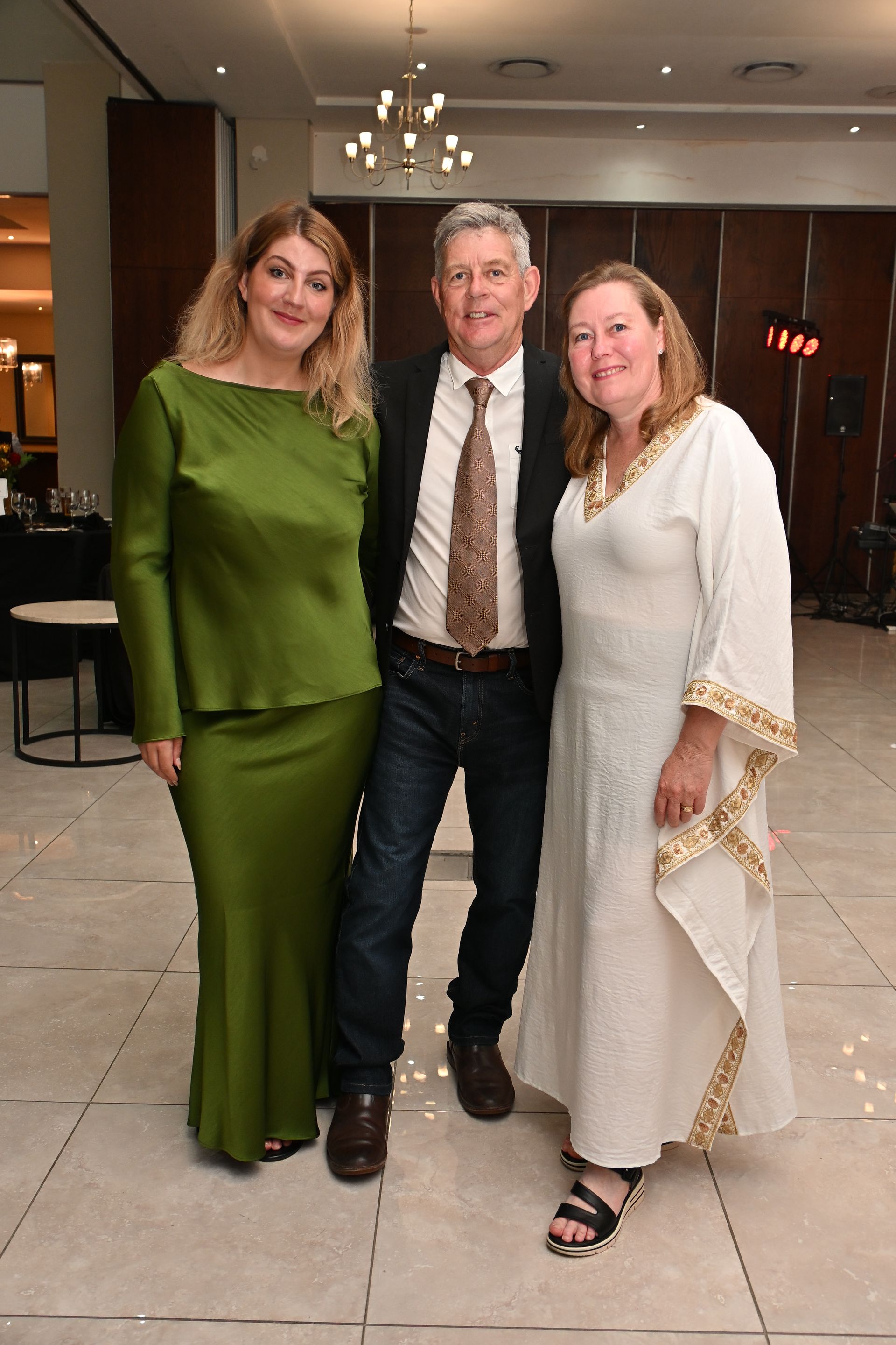 Three people posing indoors, two in cream outfits flanking one in a dark jacket and tie.
