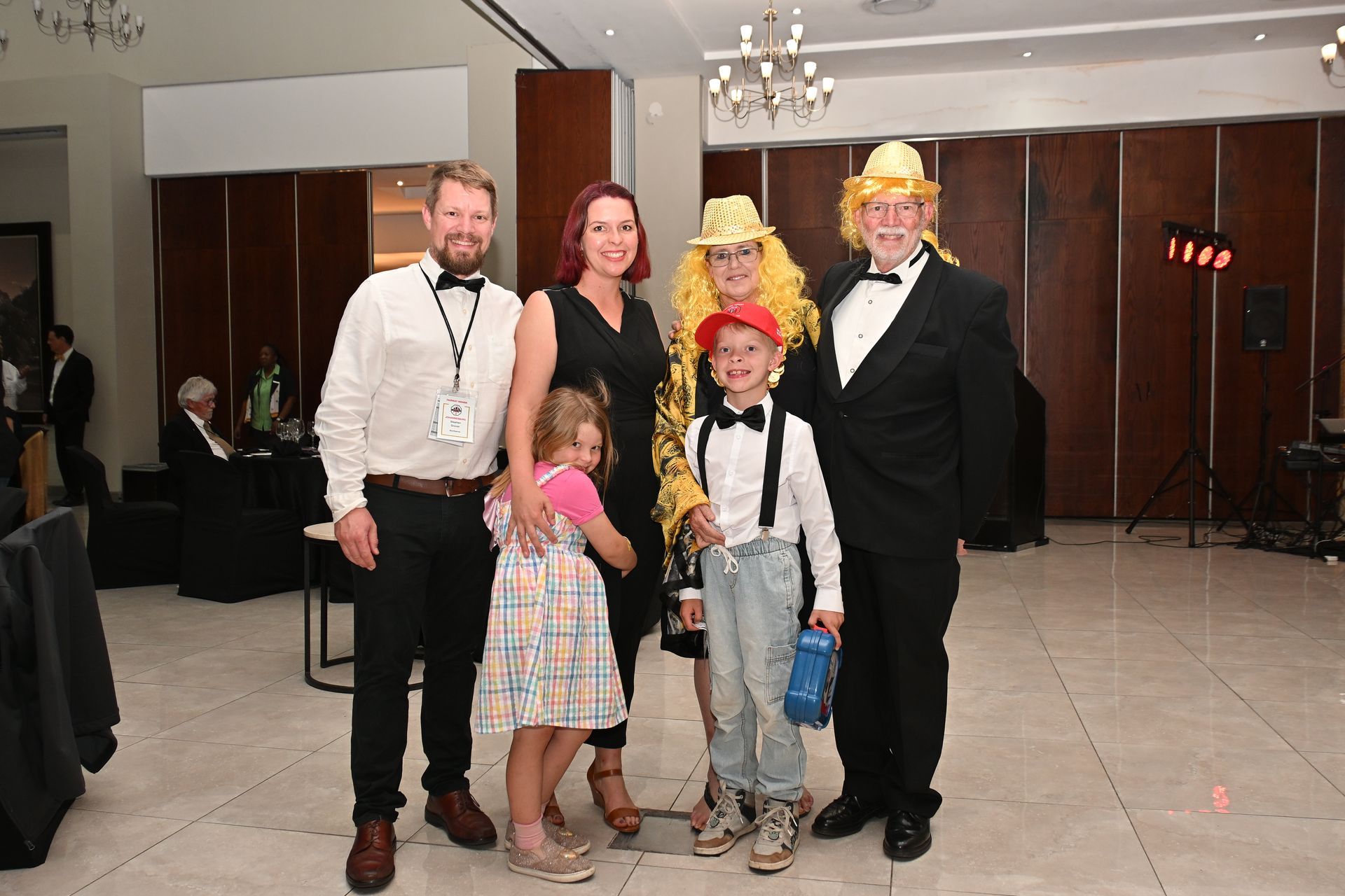 Five people posing in formal wear at an indoor event with a dark wood-paneled wall.