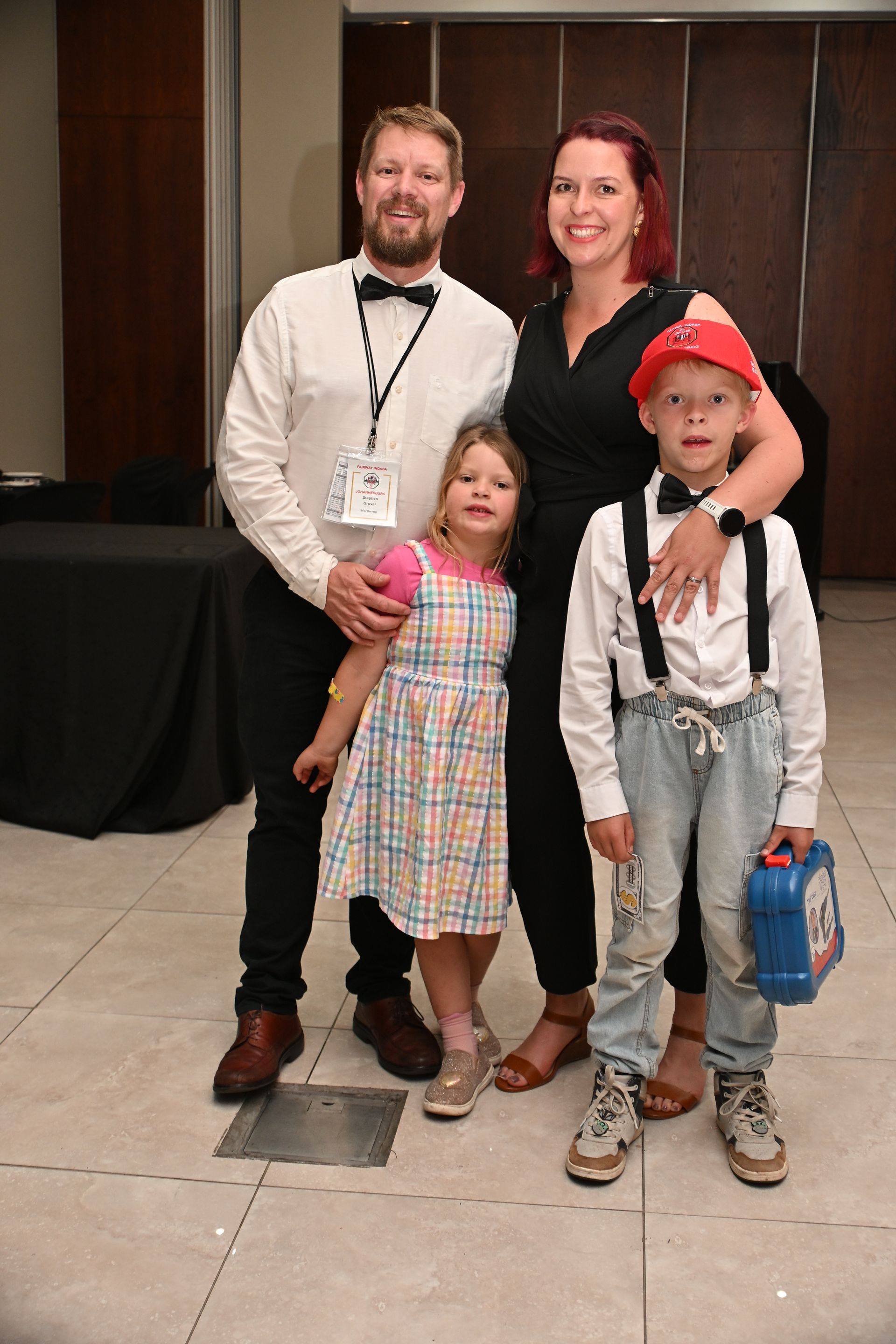 Four people posing indoors: two adults and two children, with one child holding a blue bag.