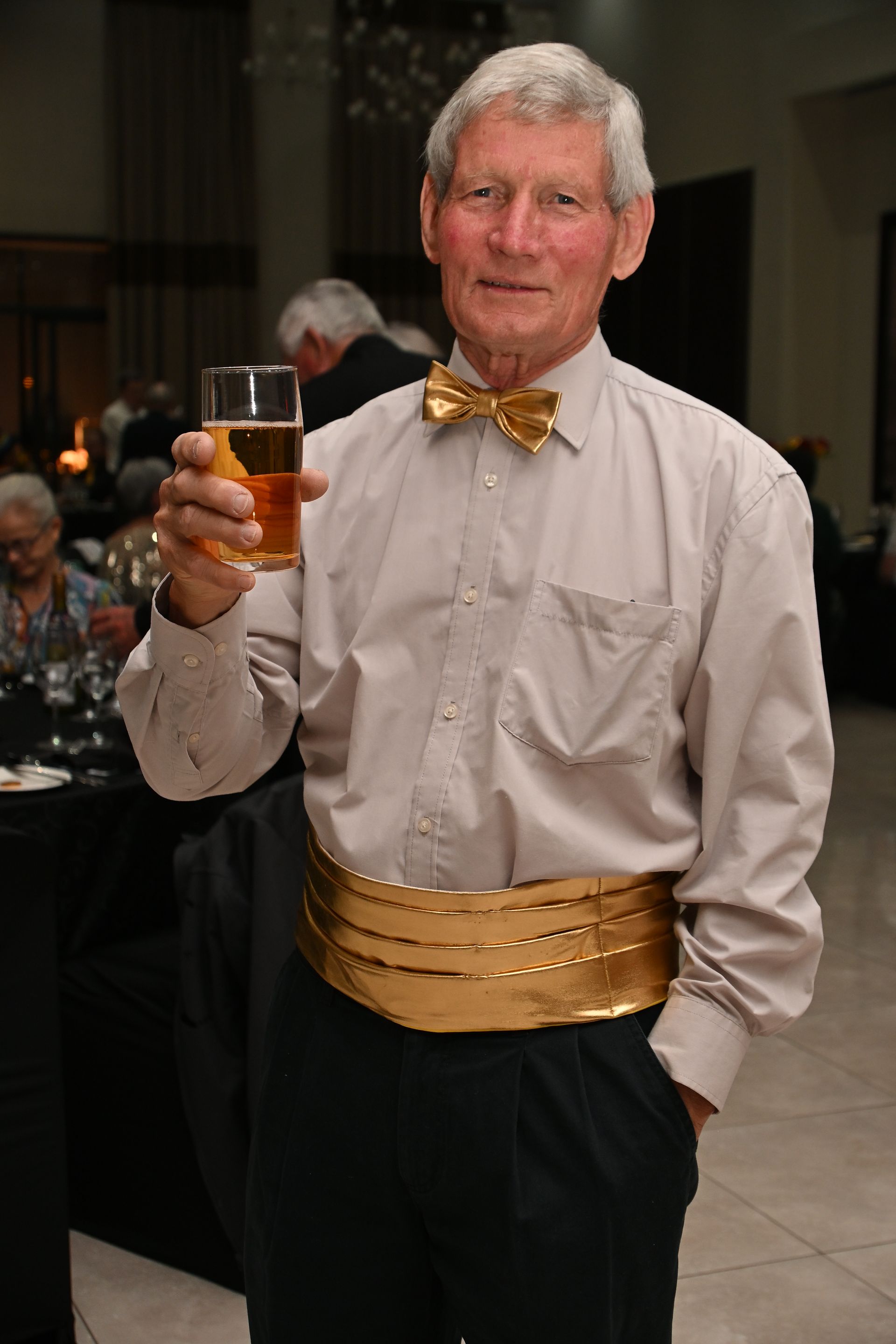 Man holding a drink at an indoor event, wearing a light shirt, bow tie, and gold sash.