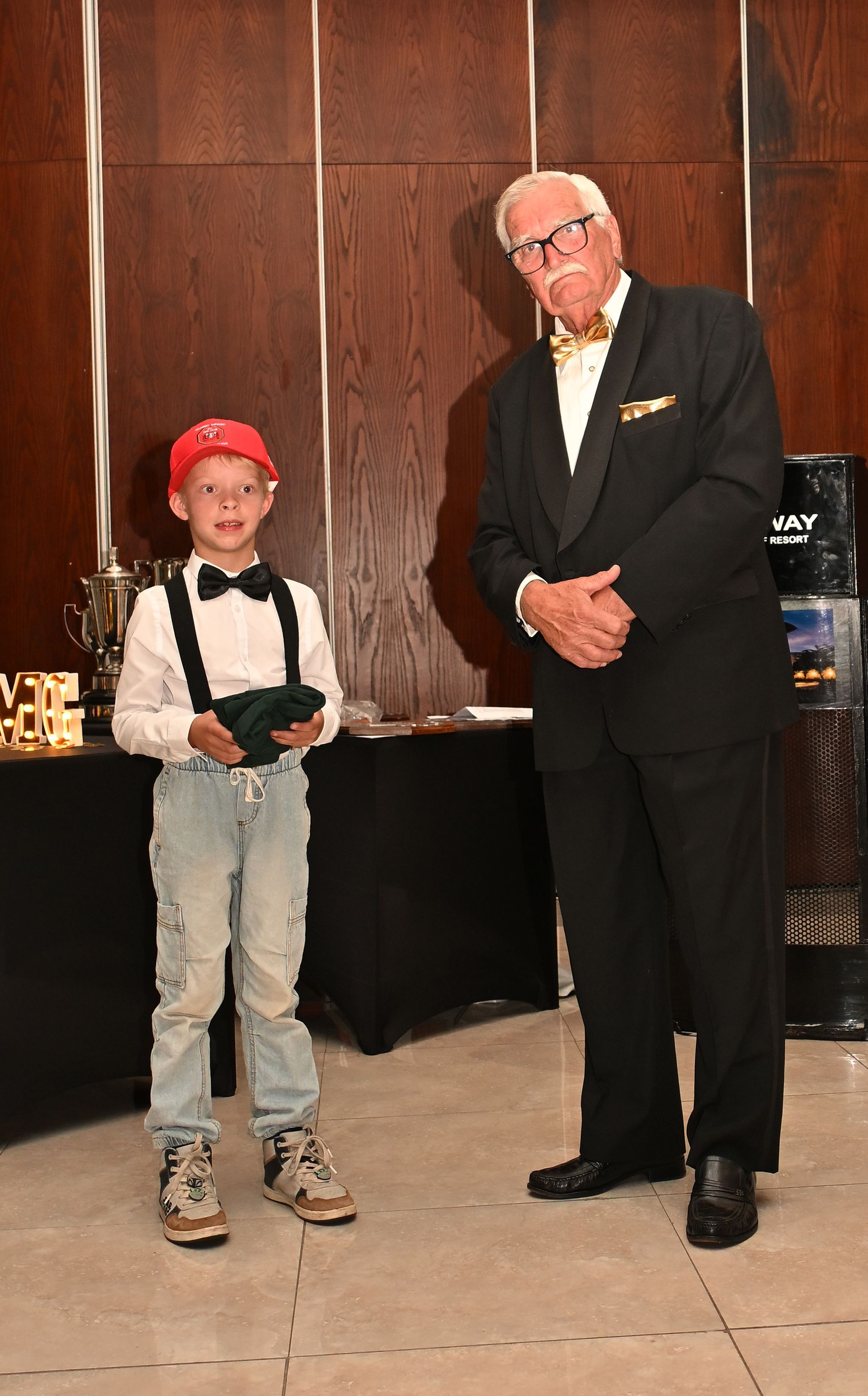 Boy in red cap standing beside a man in a suit at an indoor event