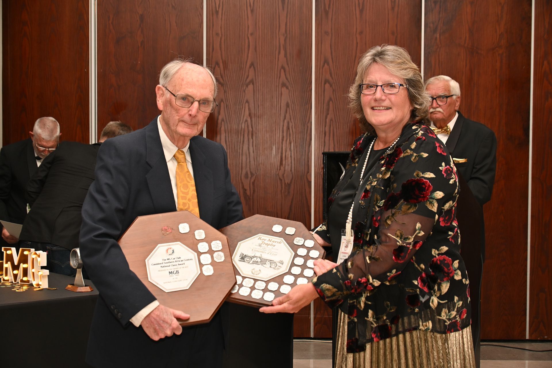 Two people holding award plaques in a wood-paneled room, with another person in the background