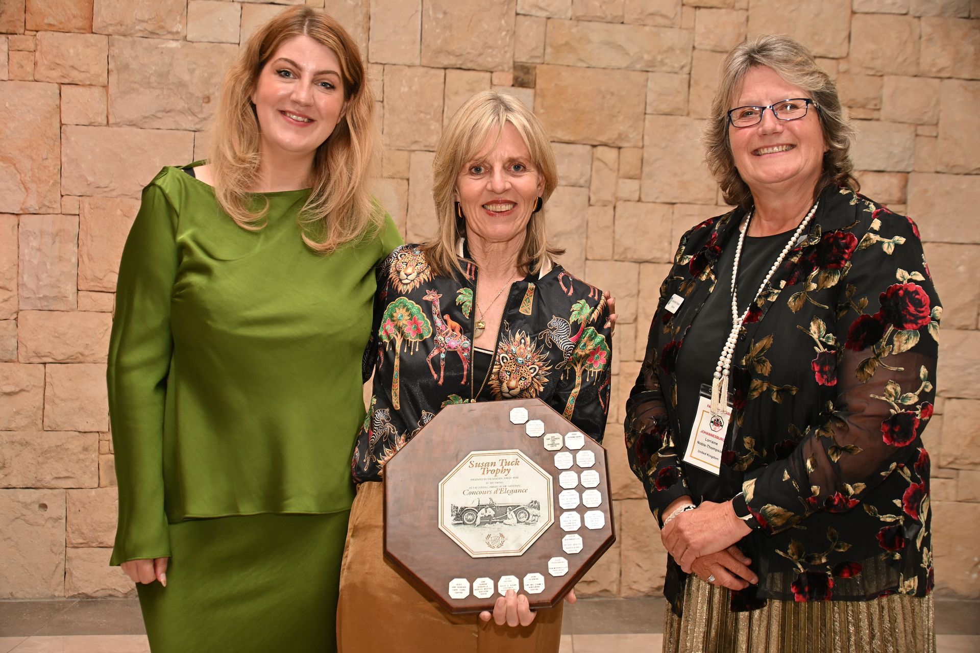 Three women posing indoors with a commemorative award plaque on a stone wall backdrop