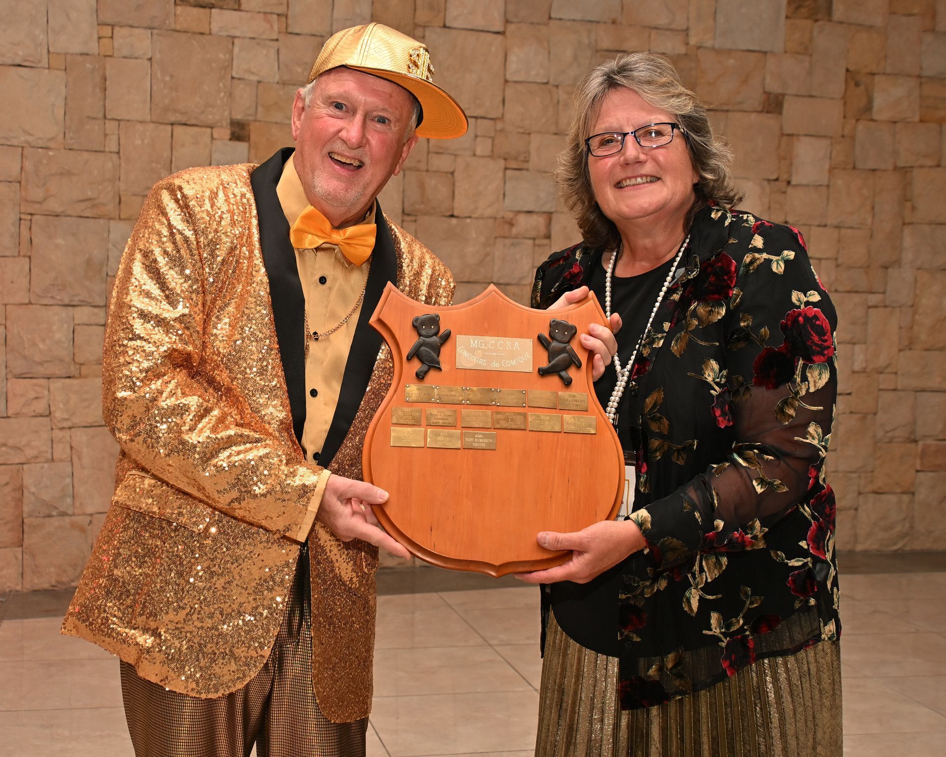 Two people smiling and holding a large orange trophy plaque against a brick wall backdrop