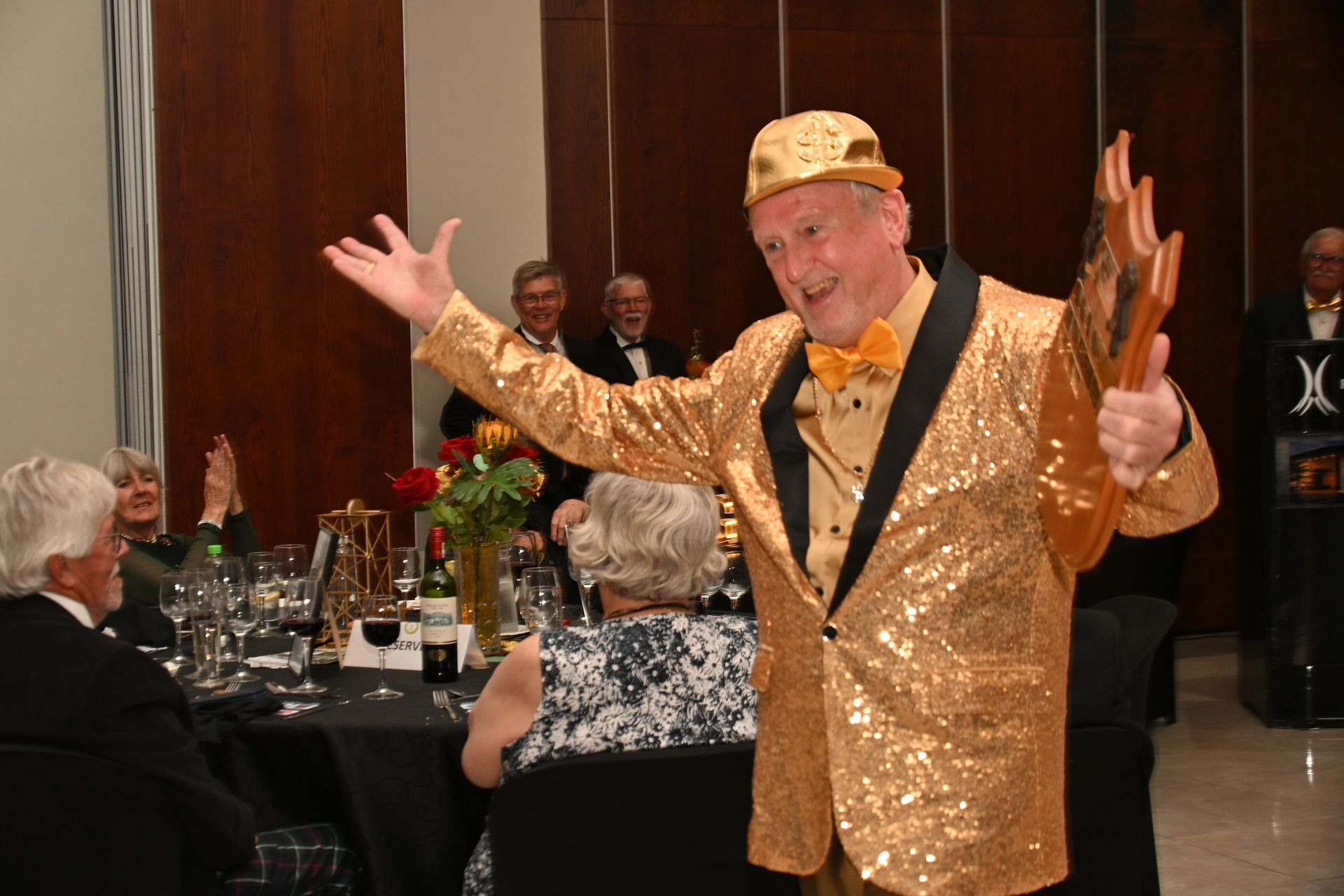 Man in a gold sequined jacket and hat posing with arms raised at a banquet table event