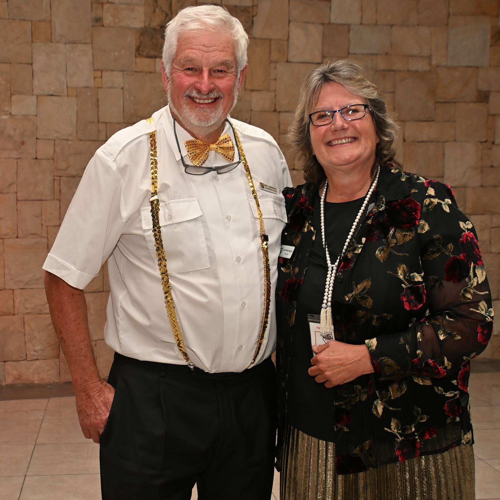 Two smiling people standing together at an indoor event, wearing formal black-and-white attire and leis.