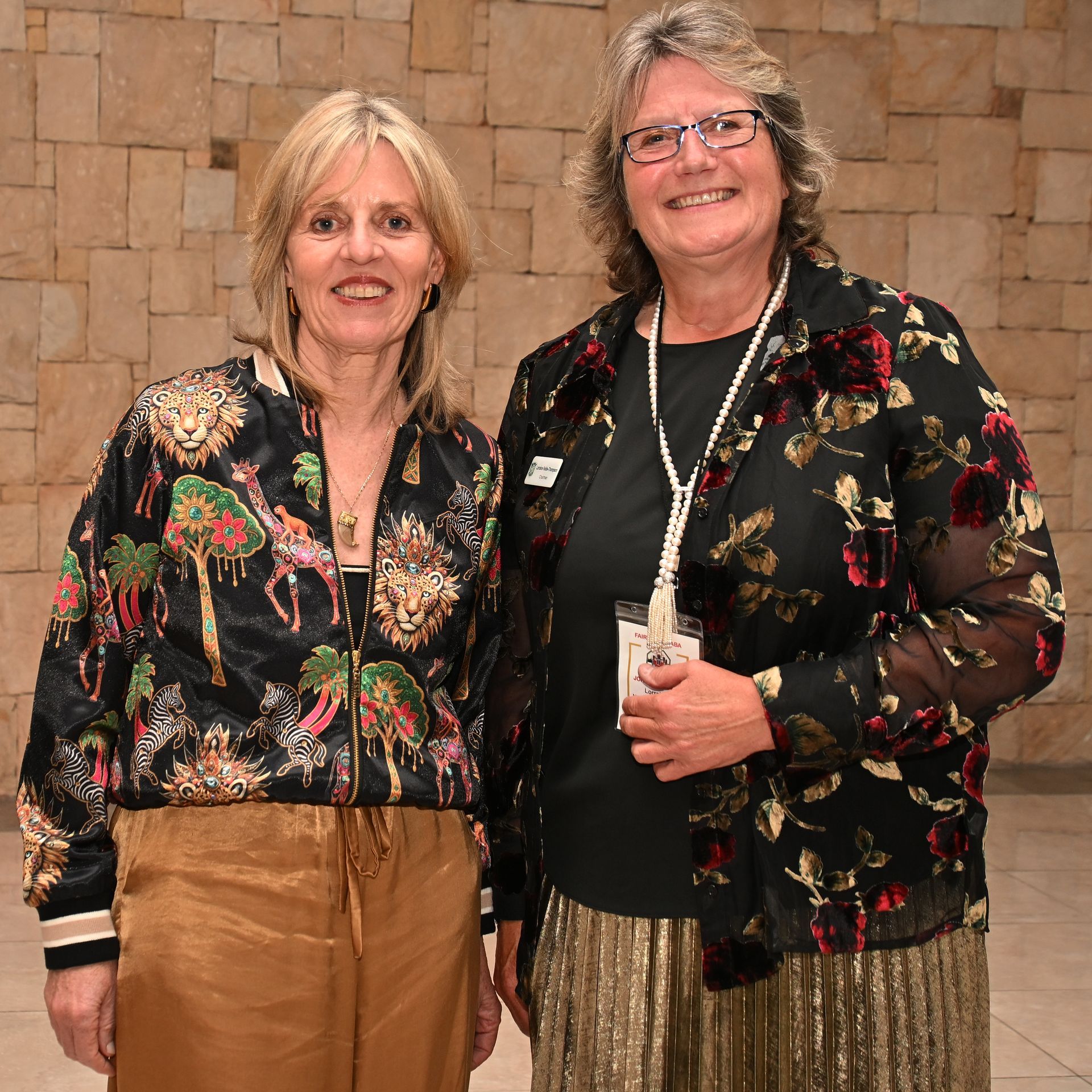 Two women posing indoors against a stone wall, wearing floral jackets and smiling.