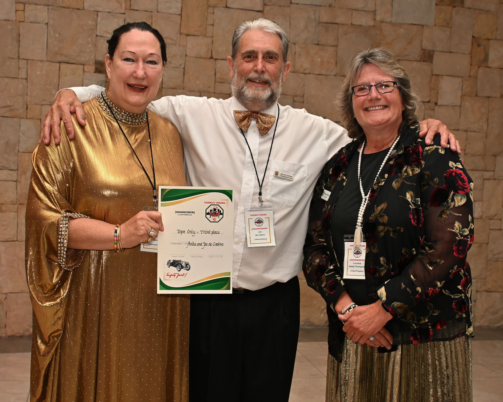 Three people posing at an event, holding an award plaque and wearing lanyards, in front of a stone wall.