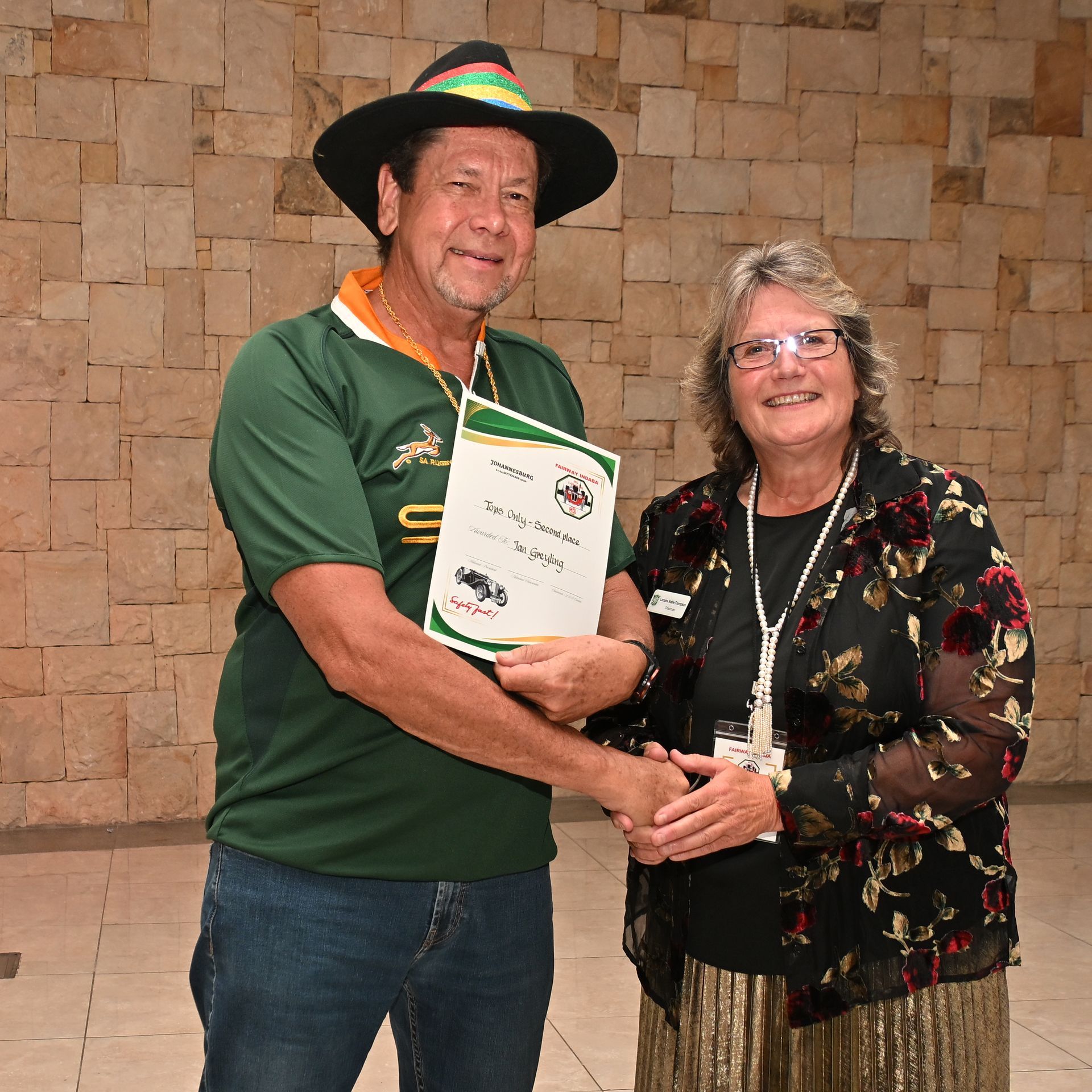 Two people smiling and holding a certificate in front of a stone wall.