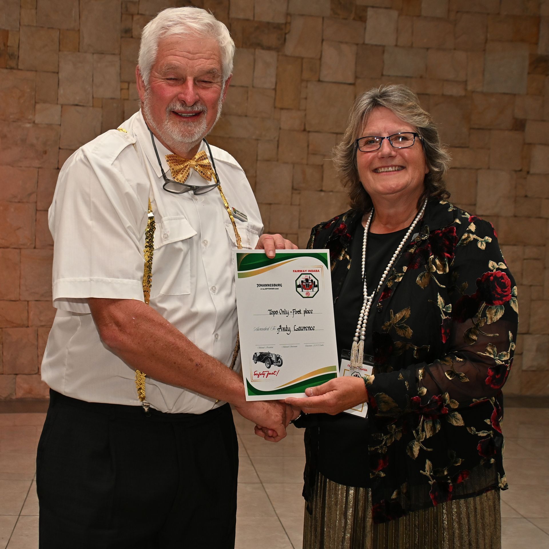 Two people hold a certificate together in front of a stone wall, both smiling.