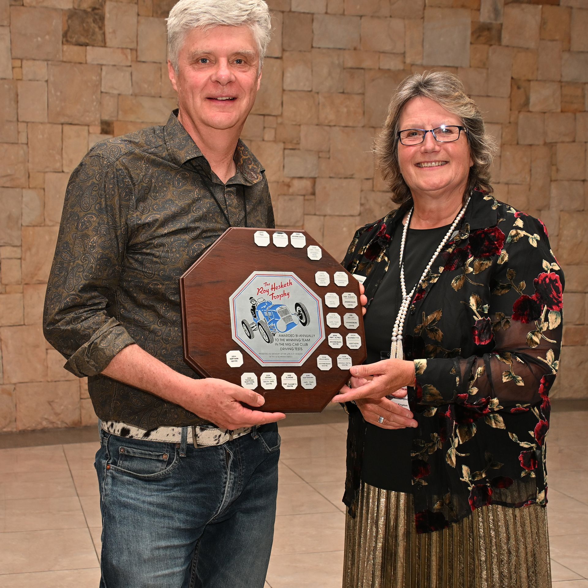 Two people holding a large wooden award plaque in front of a stone wall.