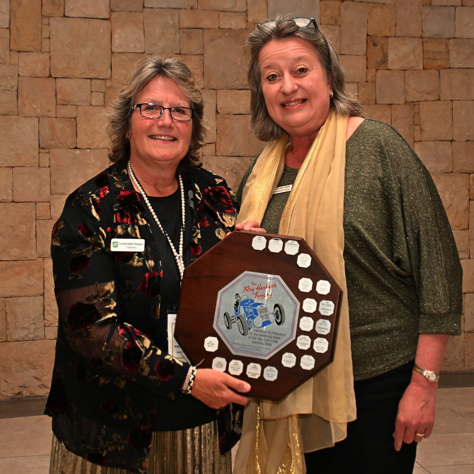 Two people smiling and holding a plaque in front of a stone wall