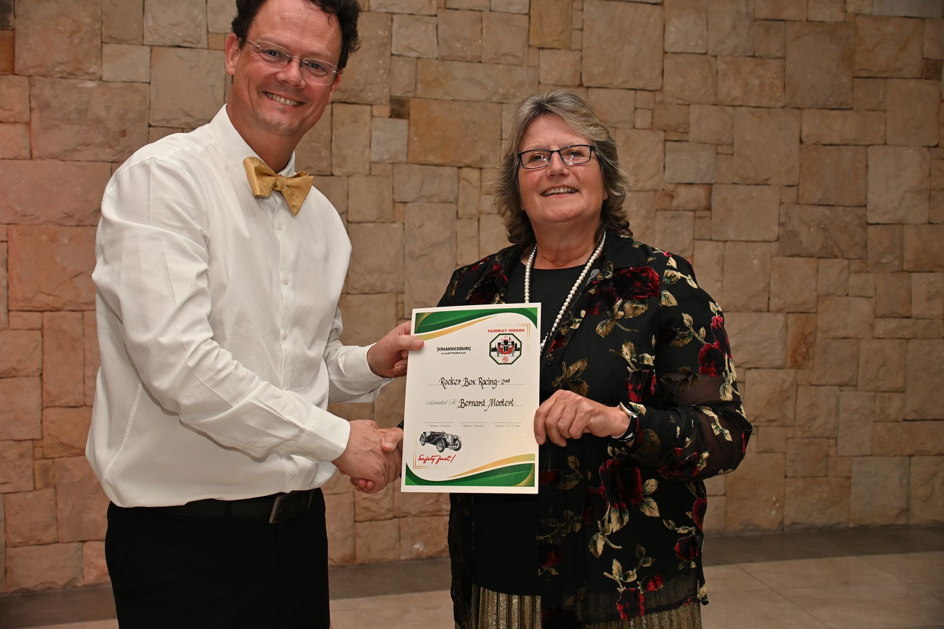 Two people holding a certificate in front of a stone wall, smiling at the camera