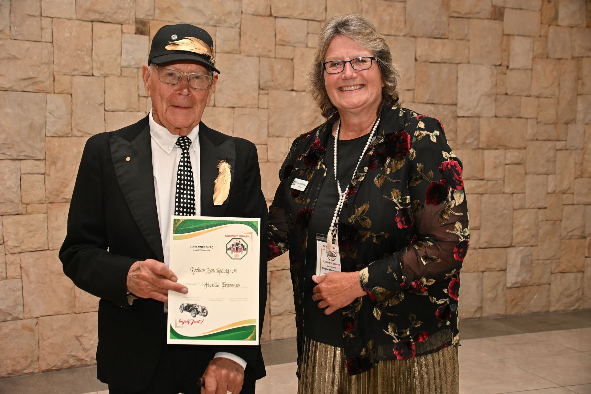 Two people standing indoors, holding a certificate and smiling against a stone wall.